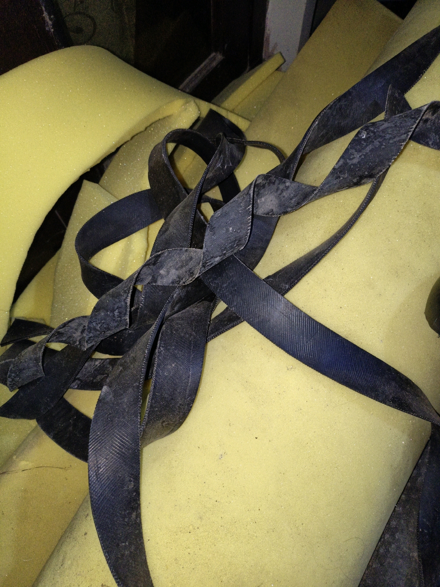 The image shows a close-up view of yellow foam material intertwined with dark, flat straps. The foam appears to be cut into various shapes and sizes, creating a layered and somewhat disorganized pile. The straps are textured with a ribbed pattern and show signs of wear, with dust and debris clinging to their surface. The lighting is dim, casting shadows and highlighting the textures of the materials. The scene appears to be indoors, possibly in a workshop or storage area. There are no people or animals visible in the image, and no specific location in Cikal, Indonesia can be identified from the visual information provided.