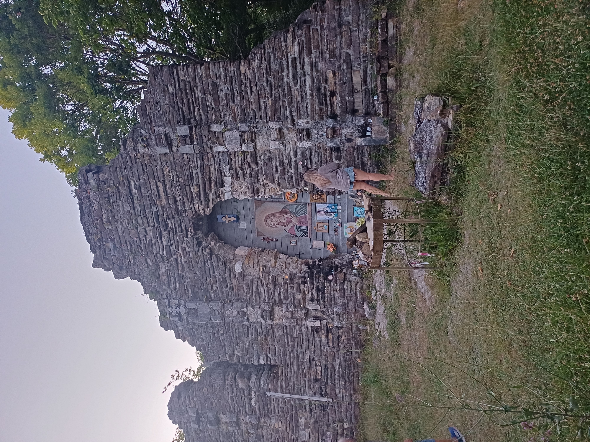 A woman stands in front of a stone structure, possibly ruins, adorned with religious icons and paintings. The structure appears to be made of rough-hewn stones stacked without mortar. A small metal table with some items sits in the foreground, and a stone fire pit is visible to the right. The area is overgrown with grass and weeds. A portion of a tree is visible at the top right. The sky is clear and pale. This location is identified as being in Golovinka, Russia.