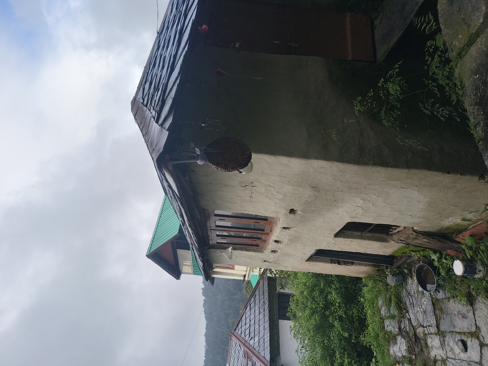 The image depicts a rustic, multi-story dwelling with a slate roof, situated in a hillside location in Haini, India. The structure is composed of textured, light-colored walls with visible imperfections. It features wooden-framed windows and a dark-colored door. Various decorative elements hang from the eaves of the house, including a circular carved item and smaller beaded ornaments. In the foreground, to the left, is a stone-paved area with overgrown vegetation. Several buckets, some containing water, are placed on the ground near the entrance of the house. Behind this dwelling, another building with a vibrant green corrugated metal roof is visible, partially obscured by the main structure. Further in the background, a dense forest covers the rolling hills under a cloudy sky, suggesting a natural, possibly damp, environment. The overall impression is one of a simple, rural homestead set against a backdrop of lush, forested terrain.