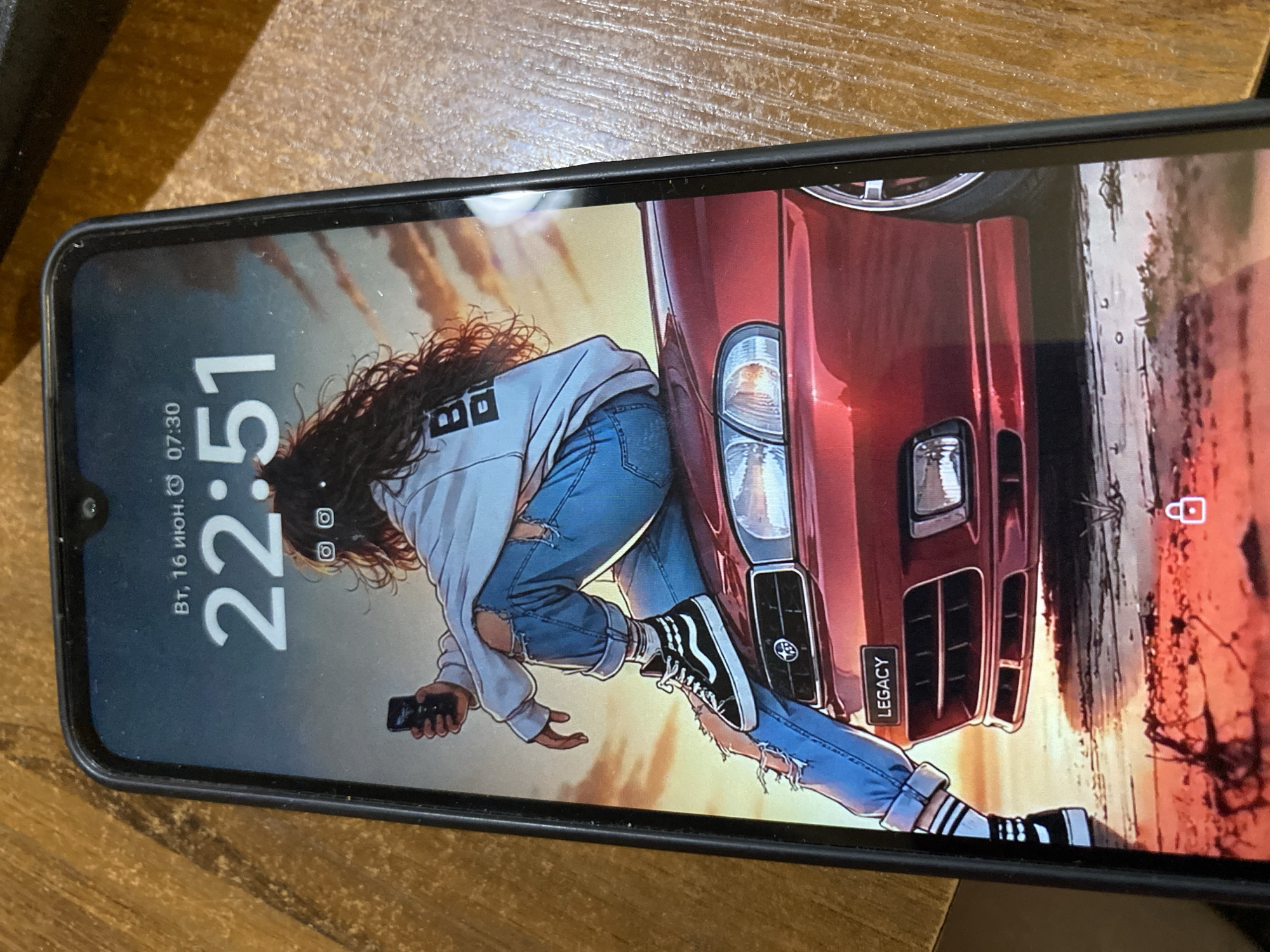 The image shows a smartphone displaying a lock screen with a digital illustration. The illustration depicts a young woman with long, dark, wavy hair, wearing a light-colored hoodie with ripped sleeves and ripped blue jeans. She is sitting on the hood of a red car, holding a smartphone in her left hand. The car appears to be a sporty sedan. The background of the illustration shows a sunset or sunrise with clouds. The time displayed on the lock screen is 22:51, and the date is "ВТ, 16 июн." (Tuesday, June 16). There are also two circular icons with dots inside below the time. The bottom of the screen shows a padlock icon. The smartphone itself is black and is placed on a wooden surface, possibly a table. There is no indication of the city of Almaty, Kazakhstan, or any specific urban environment in the image.