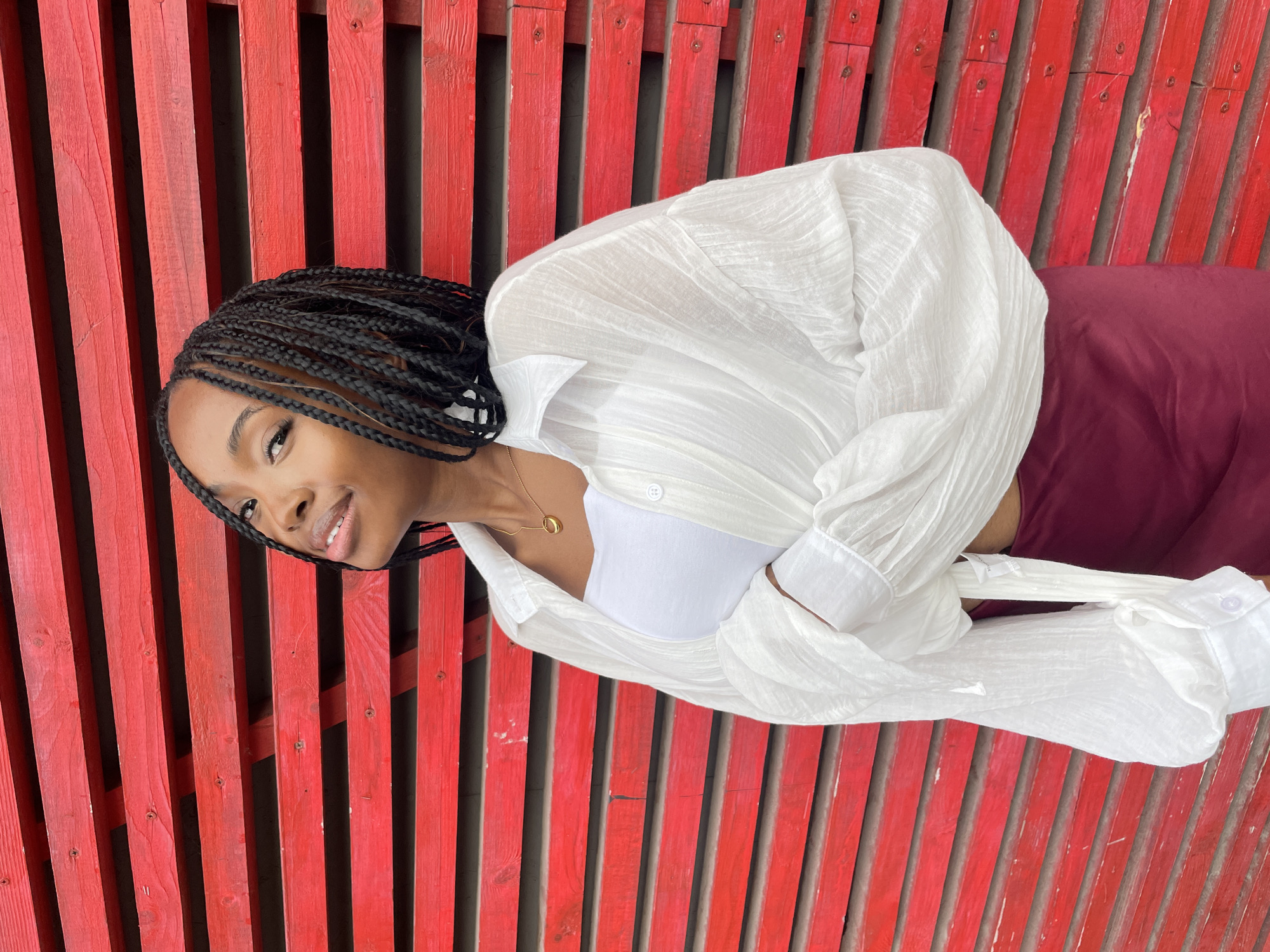 A young Black woman with braided hair and a white shirt stands in front of a red wooden wall. She is wearing a white tube top and a burgundy skirt. She is looking up and to her left, with a slight smile on her face. The red wall is made of horizontal wooden planks.