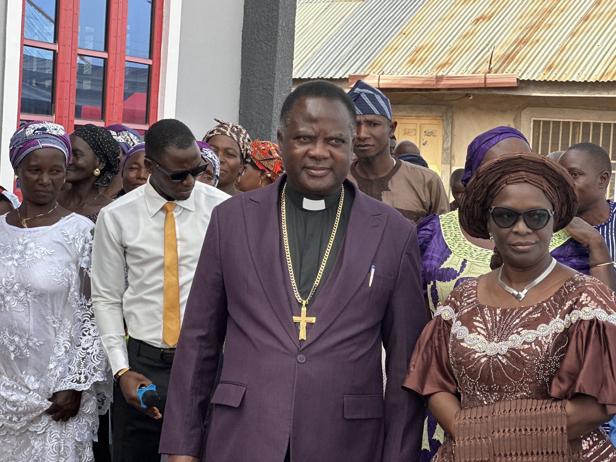 The image captures a group of people standing outdoors. In the foreground, a man in a purple suit with a clerical collar and a gold cross necklace is the focal point. He has a pen in his pocket. To his left is a man in a white shirt and yellow tie, wearing sunglasses and holding a blue microphone. Further to the left is a woman wearing a white lace dress and a patterned head covering. To the right of the man in the purple suit is a woman wearing a brown patterned dress and sunglasses, also with a patterned head covering. Behind the foreground figures, there are several other people of varying ages and attire, many with head coverings. The background shows a building with red-framed windows to the left, and a corrugated metal roof to the right. The location is Goni Gara, Nigeria.