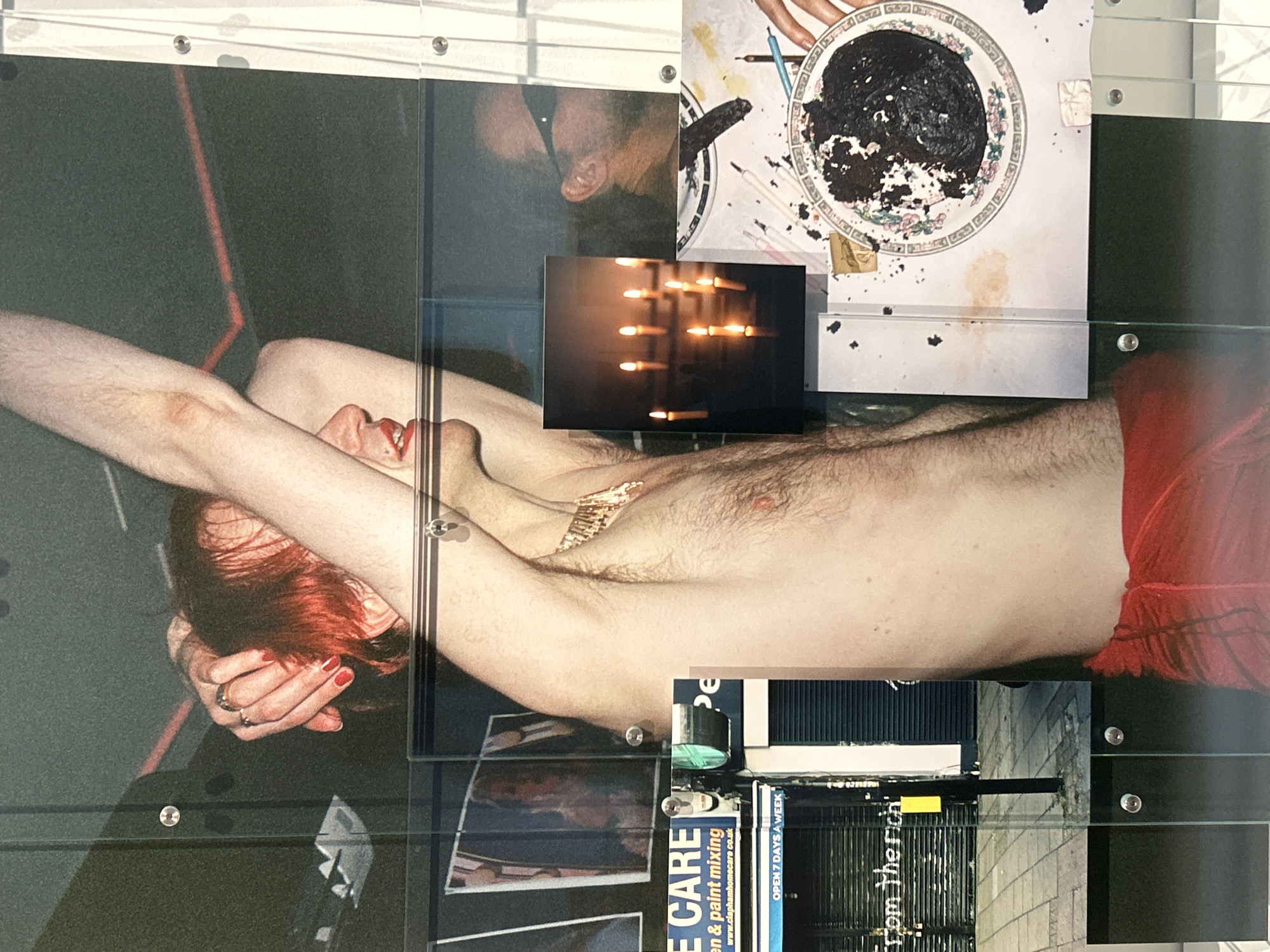 The image is a layered collage of photographs and prints affixed to a transparent surface, possibly acrylic or glass, held together by bolts.
A central, horizontally-oriented photograph depicts a pale-skinned person with red hair, lying on their back, arms crossed over their head, and wearing red shorts. A gold necklace is visible.
Overlaid on this are several smaller images. To the left, a photograph shows two faces close together. In the upper right, a plate smeared with black material is depicted. Below, a photograph shows lit candles, and another reveals a street scene, potentially with a shopfront or building façade.
The street scene includes the words "E CARE" and mentions "paint mixing." It also mentions "www.claphamhomecare co.uk" and "OPEN 7 DAYS A WEEK," suggesting a shop or business in Clapham, London, United Kingdom. The words "from the rich" are also visible.