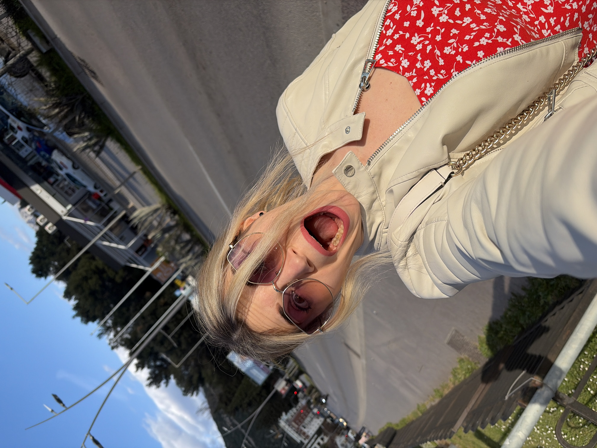 An outdoor, tilted selfie captures a blonde-haired female subject from the chest up, positioned on a roadside in Budva, Montenegro. She wears large, pink-tinted, square-framed sunglasses and a light beige leather-style jacket over a red garment with a white floral pattern. Her mouth is wide open, revealing her teeth, in an expressive manner. A gold-colored chain strap from a bag is visible over her right shoulder.
The background shows a two-lane asphalt road, identified as Jadranski put, with traffic visible in the distance. On the right side of the road, commercial buildings are present, notably a white structure with a red awning, consistent with a Jugopetrol gas station (specifically the one at the top of the hill on Jadranski put). Beyond the road and buildings, dense green trees cover what appears to be a hillside or mountain. Modern streetlights with thin poles extend into the clear blue sky, which contains scattered white clouds. Sunlight illuminates the scene, casting shadows, indicating a bright day. A metal fence and grassy verge are visible in the foreground to the left.