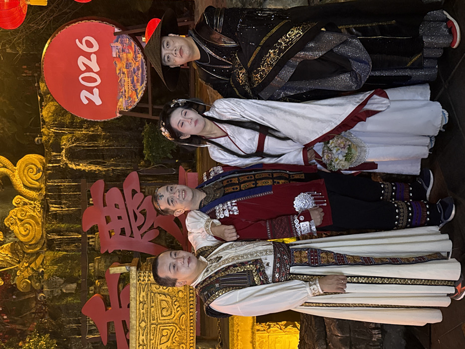 Four people in traditional Chinese costumes pose for a picture in front of a decorative wall. The first is wearing a black outfit and a hat. The second, a white dress and holding flowers, has her eyes closed. The third is wearing a red and black outfit. The fourth, wearing an ornate white and gold outfit, is smiling. A red circle with the number 2026 on it is visible above the group. The background includes ornate decorations.