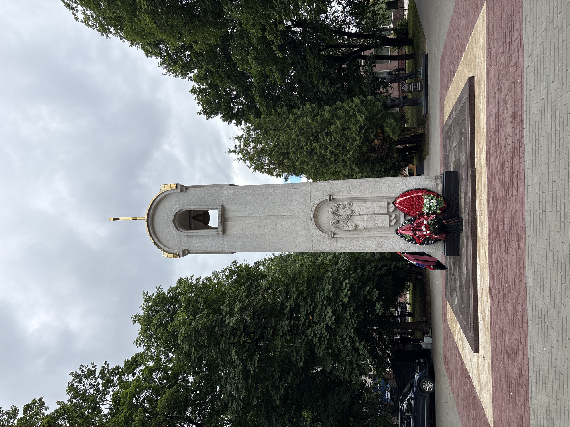 The image shows a war memorial located in the city of Radkevshchina, Russia. The memorial is a tall, light-colored structure topped with a gold-colored cupola and cross. A bell is visible within an arched opening near the top. The front of the memorial features two carved figures in relief, appearing to be religious or historical figures. At the base of the memorial, two wreaths of red flowers and greenery are placed. The memorial is situated in a paved plaza with patterned brickwork. In the background, several large, mature trees with dense green foliage line the area. To the left of the memorial, a dark-colored SUV is parked. In the further background, there are indications of buildings and other structures, along with more trees and what appear to be statues. The sky is overcast and cloudy, suggesting a dull day. The overall scene depicts a solemn and commemorative outdoor space.