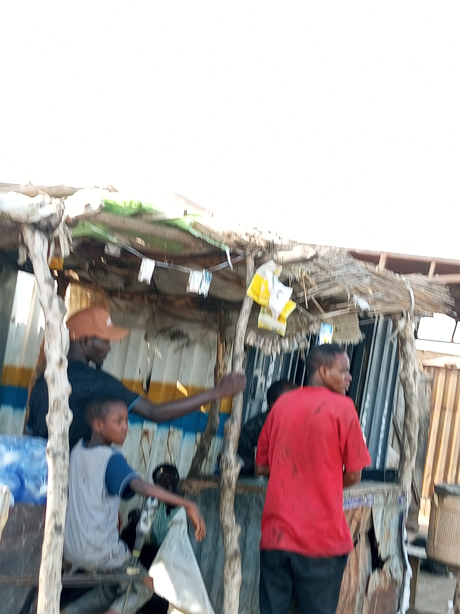 A low-angle, medium shot captures a group of people gathered around a makeshift structure, possibly a roadside stall, in Gadao, Nigeria. The structure is constructed from corrugated metal and wood, with a thatched roof. In the foreground, a young boy in a grey and blue t-shirt sits on a wooden bench, looking to his right. To his left and slightly behind him, an adult male wearing a dark shirt and an orange baseball cap is visible, his arm extended as if reaching for something. To the right of the frame, a person in a red t-shirt and dark pants stands facing away from the camera, their back to the viewer. Further back, another person in a dark shirt is partially visible. The background consists of the rough, weathered walls of the stall and some hanging items. The overall scene depicts a casual gathering in an outdoor, informal setting.