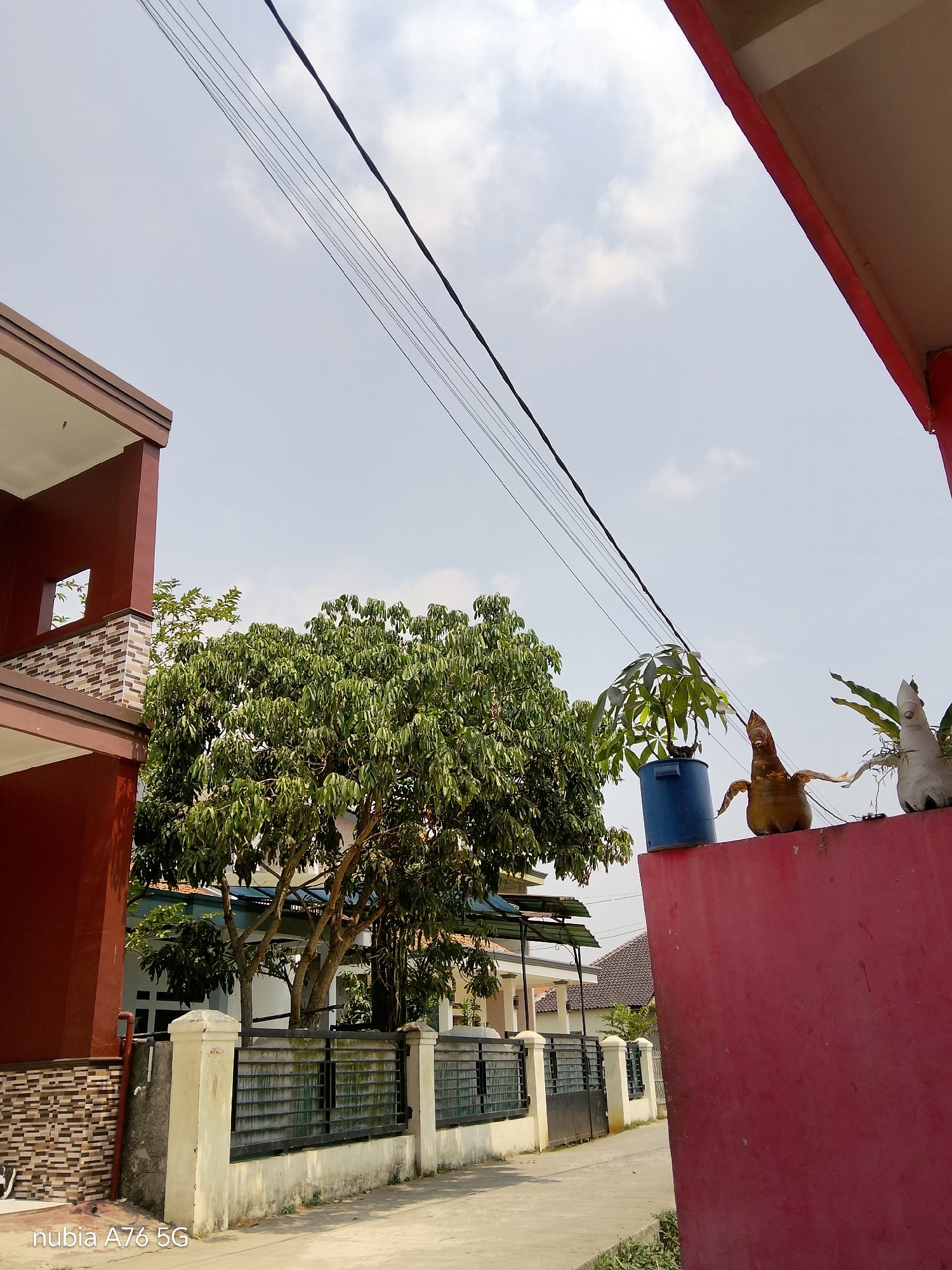 The image depicts an outdoor scene in Cakung, Indonesia. In the foreground, a red wall dominates the right side, with decorative objects and potted plants positioned on its top edge. To the left of the wall, a street is visible, bordered by a fence and a cream-colored wall. Beyond the fence, a large tree with dense green foliage stands prominently, with a multi-story building situated to its left. Another building, characterized by its reddish-brown exterior and patterned tiling, is also visible on the far left. Overhead, several power lines stretch across a sky with scattered clouds. The lighting suggests it is daytime.