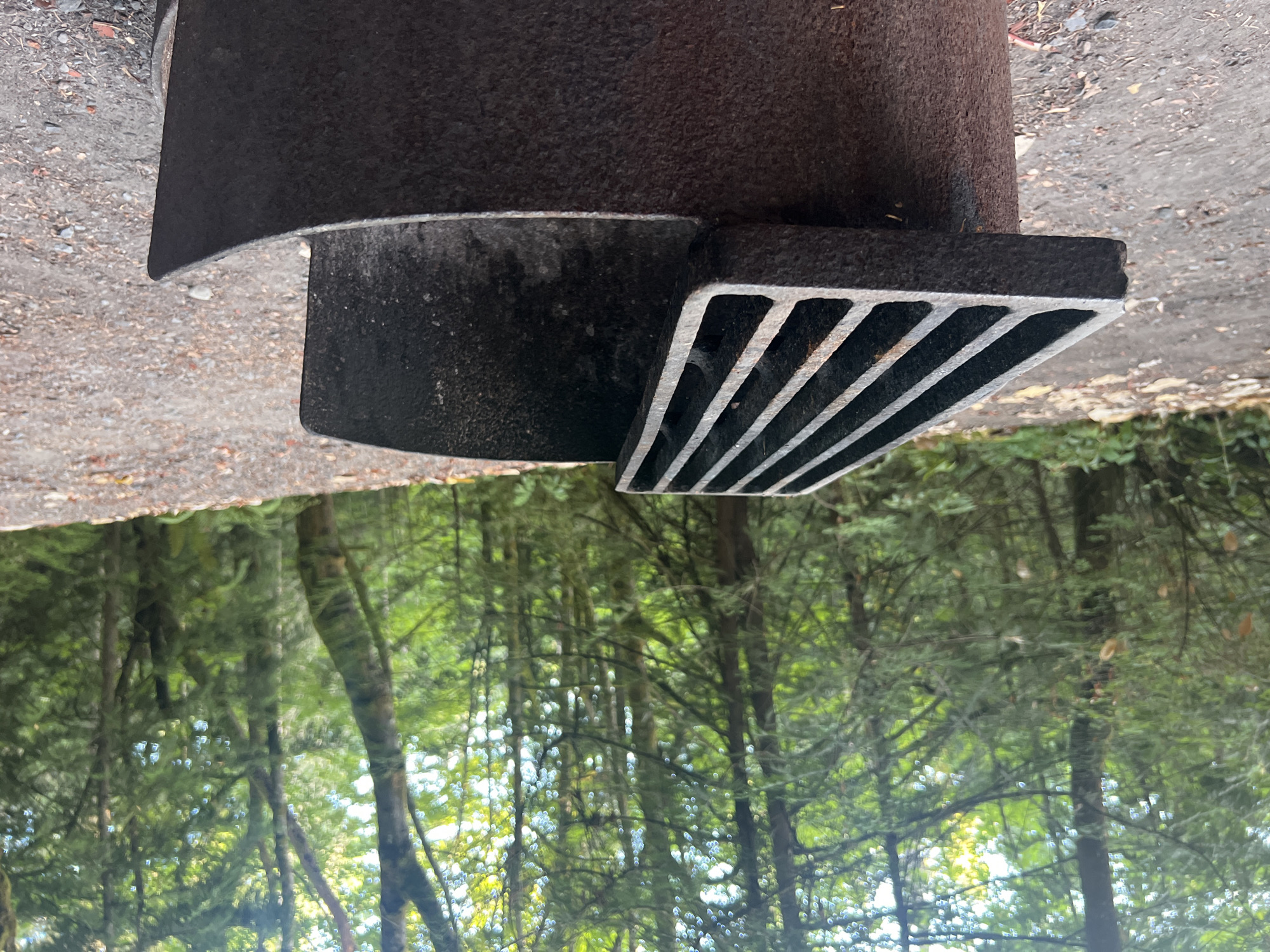 A close-up, eye-level, medium shot shows a rusty, weathered metal fire pit in a forested area. The fire pit's cylindrical base is partially visible, supporting a heavy, slatted metal grill. The grill is tilted, with its right side resting on the rim of the pit and its left side angled upwards. The background is a dense forest with tall trees, their trunks dark and textured, and the canopy a vibrant green, dappled with sunlight. The ground in the foreground is a mixture of gravel and dirt, with fallen leaves and small stones scattered around. The scene appears to be located in a park or campground within Area A (Mill Bay / Malahat), Canada. There are no people or animals visible in the image.