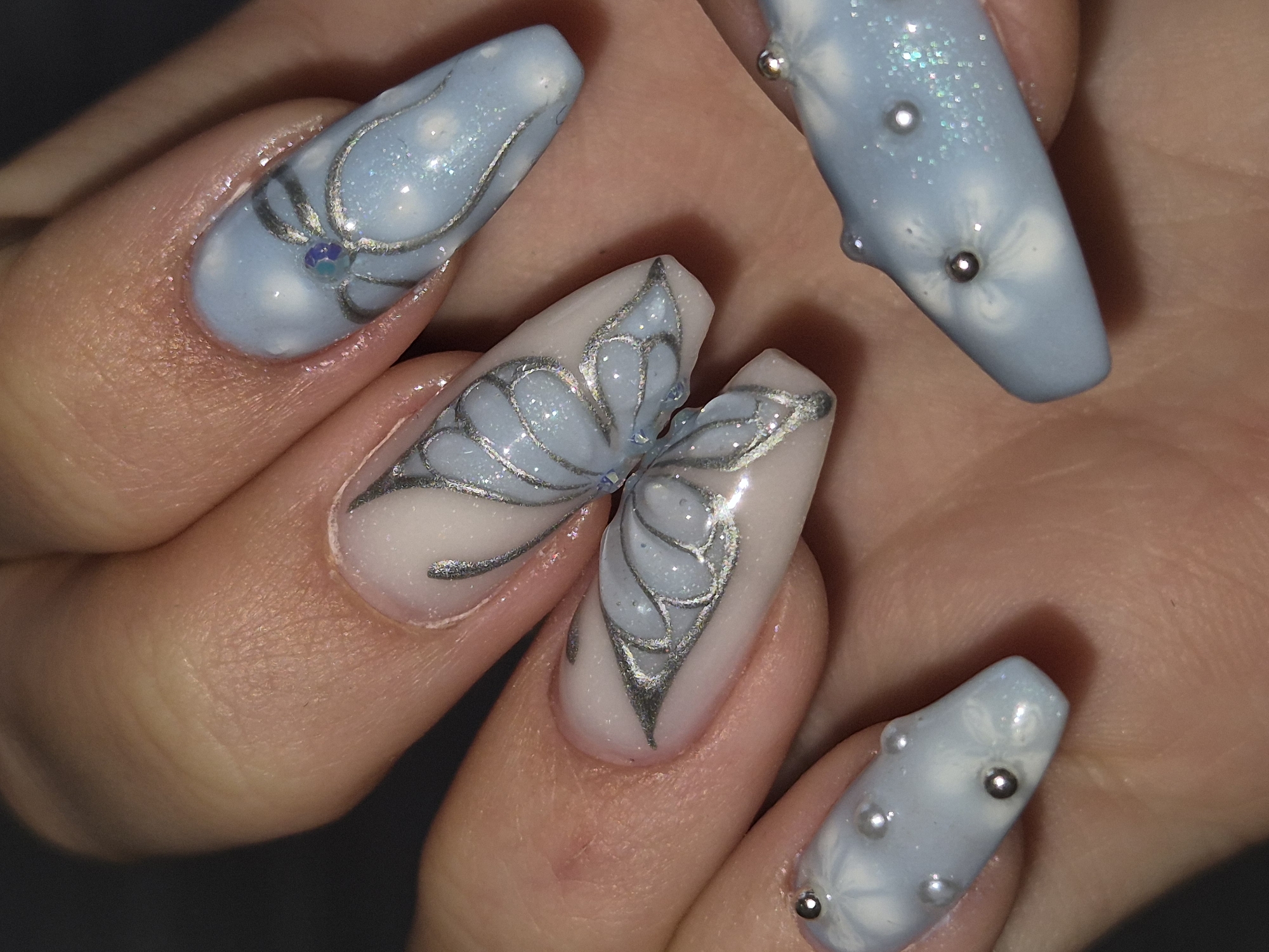 The image displays a close-up, high-angle view of an adorned hand, likely showcasing intricate nail art in Pavlikeni, Bulgaria. The subject's hand is visible, featuring five long, artificial nails with elaborate designs.
Three nails are prominently in view: the index finger, middle finger, and ring finger. The index finger and pinky finger nails are a soft, glossy light blue, embellished with delicate, matte white floral patterns that appear to be sculpted or painted in a 3D style. These blue nails are further decorated with small, shiny silver metallic beads and scattered iridescent glitter, some of which are tiny rhinestones.
The middle and ring finger nails combine to form a large, stylized butterfly. The background of these nails is a creamy off-white or very pale pink, over which a shimmering silver-grey outline defines the butterfly's wings. The inner sections of the butterfly wings are filled with a translucent, light blue polish, heavily infused with fine glitter, creating a sparkling effect. The butterfly's body is subtly suggested at the base of the middle finger nail, with an accent of a tiny blue rhinestone or bead.
The nails are long and appear to be of a coffin or tapered square shape. The skin around the nails is smooth with natural creases. The lighting is bright, emphasizing the reflective qualities of the glitter and metallic beads. There is no visible text. The image captures a detailed example of artistic nail enhancement.