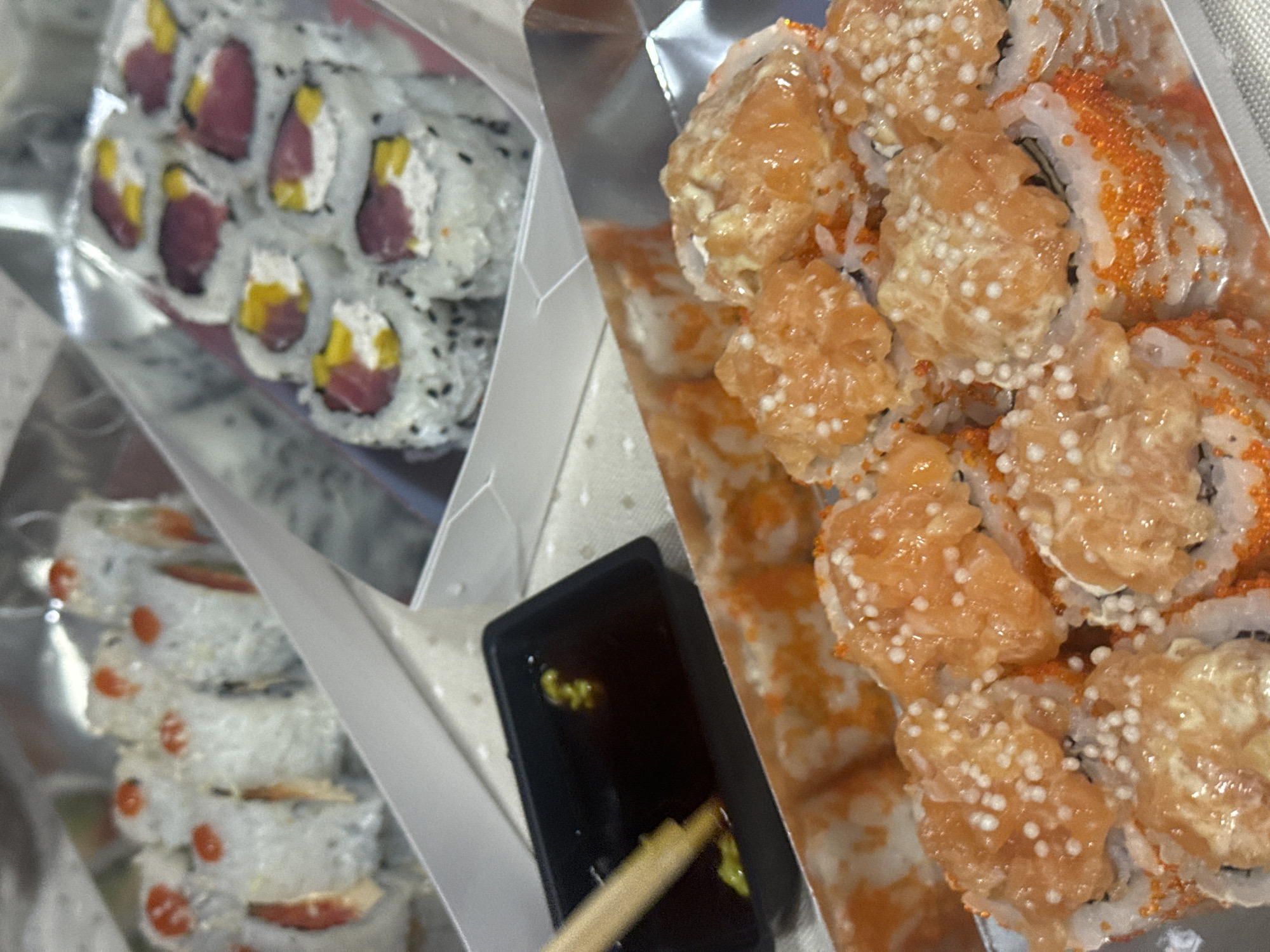 The image displays a close-up, overhead view of a sushi meal, likely prepared for take-out or delivery, set in Krasnoyarsk, Russia. The scene features multiple types of sushi rolls, a container of soy sauce, and a pair of chopsticks.
In the top left, a container holds several inside-out sushi rolls (uramaki) featuring white rice coated with black sesame seeds. The visible fillings include dark red slices, likely tuna, and bright yellow pieces, possibly mango or pineapple, creating a colorful contrast.
To the right, a separate container is filled with more elaborately topped sushi rolls. These rolls are adorned with numerous small, bright orange roe (tobiko or masago) and appear to have a generous topping of what looks like finely minced or sauced salmon, mixed with small white spherical textures, possibly tempura flakes or another crunchy element.
Partially visible in the bottom left, another stack of sushi rolls can be seen, characterized by white rice and small red toppings, though specific details are less clear due to blurring and partial obstruction.
In the foreground, a small, black rectangular dish holds dark soy sauce with two distinct dollops of green wasabi. A wooden chopstick is positioned, dipping into the soy sauce, indicating that someone is about to eat. The sushi is presented in disposable, foil-lined containers, suggesting a casual dining experience at home or another informal setting. The lighting is artificial, consistent with an indoor environment. There is no visible text in the image.