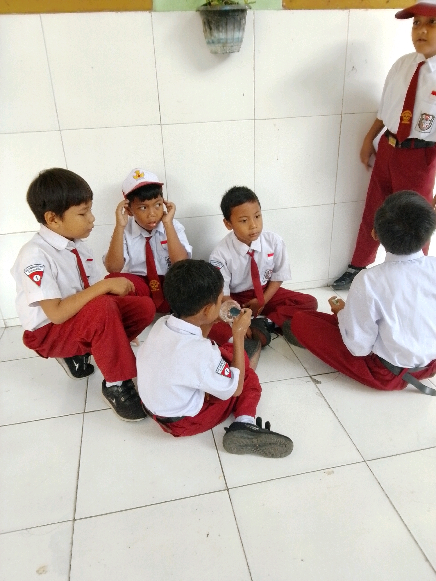 Six young boys, dressed in school uniforms consisting of white shirts with red ties and red trousers or skirts, are gathered on a tiled floor indoors. Five boys are seated in a group, facing each other or looking away, while one boy stands in the background. The boy in the background wears a red cap and a white shirt with a red tie, and red trousers. One of the seated boys holds a small dark container. The environment appears to be a school setting, possibly a hallway or classroom, with white tiled walls. The lighting suggests an indoor, possibly daytime scene. The image is taken from a high angle, looking down on the children.
