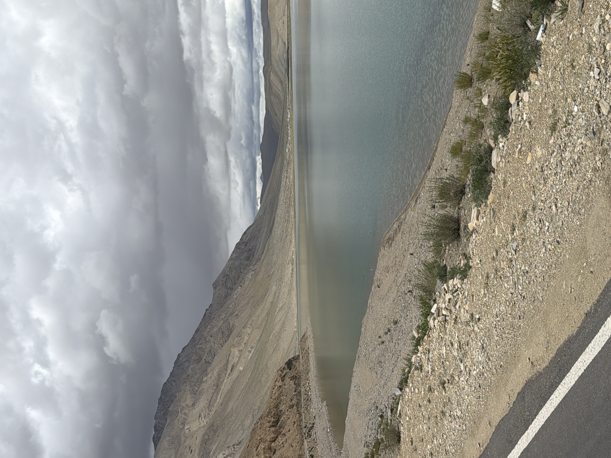 This is a landscape photograph of a serene lake and a mountainous region under a cloudy sky. In the foreground, a section of a paved road with a white stripe is visible, bordering a rocky, barren shoreline. The shoreline is sparsely populated with small, dry shrubs and scattered rocks.
The lake itself occupies the majority of the midground and extends into the background. The water appears calm and reflects the muted tones of the sky and surrounding landscape. The color of the water ranges from a muted blue-green to a lighter, almost silvery hue.
In the background, a large, imposing mountain dominates the left side of the frame, its slopes barren and streaked with erosion patterns. The mountain is a sandy, beige color. Further in the distance, a range of softer, rolling hills stretches across the horizon, with a small settlement visible nestled at their base. The sky is overcast with thick, grey clouds, suggesting a potentially cool or damp climate.
The location is identified as the city of Karzok, India. Karzok is a village situated on the shore of Tso Moriri lake in Ladakh, India. The scene depicted aligns with the typical landscape of this high-altitude region, characterized by its vast lakes, rugged mountains, and arid terrain. There are no people or specific actions visible in the image; it is a static depiction of the natural environment.