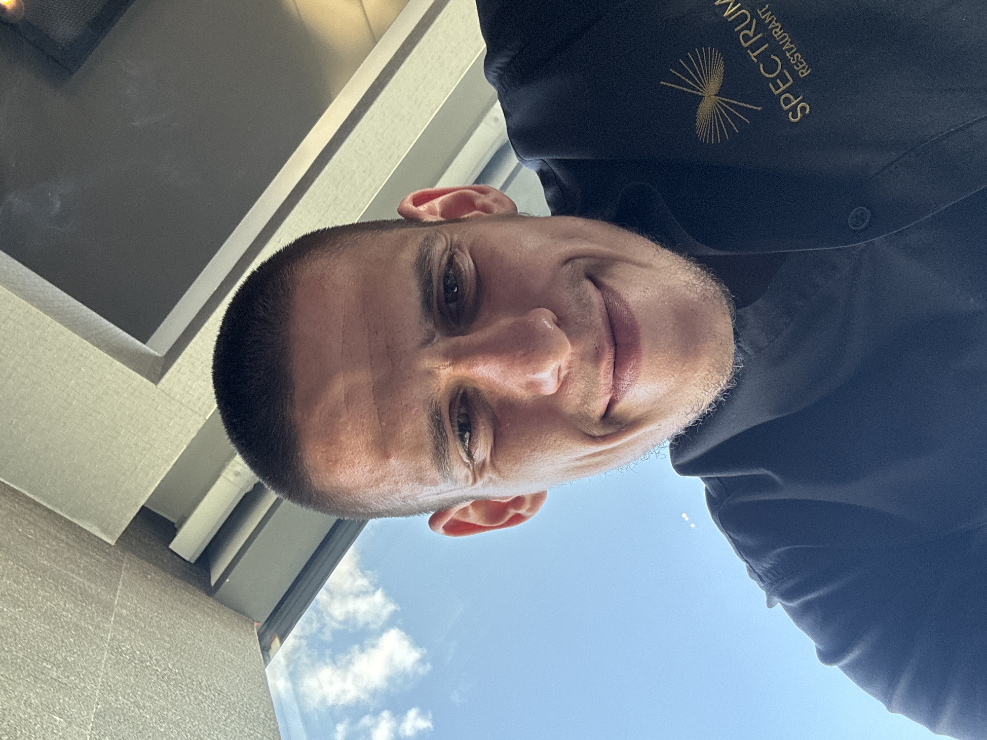 A low-angle selfie is taken by a man with a buzz cut, looking upwards towards the camera. He is wearing a dark blue collared shirt with a gold embroidered logo on the right side that reads "SPECTRUM RESTAURANT". The background consists of a bright blue sky with scattered white clouds, and the underside of a building or structure with beige and grey panels. The man has a light beard stubble and is smiling. The lighting suggests it is daytime. There are no visible indications of the city of Sofia, Bulgaria.