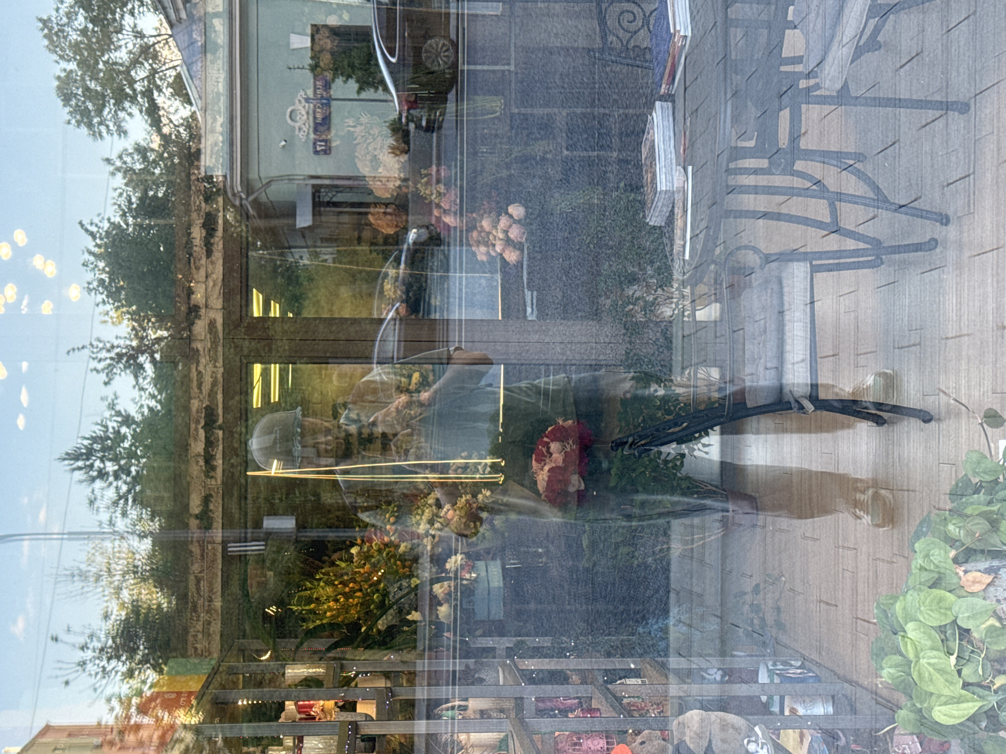 This image is a reflection captured in a large glass shopfront window on Spiridonovka Street in Moscow, Russia, during daylight hours. The reflection overlays the interior of a flower shop with the exterior urban environment.
Centrally, a person, identified as the photographer, is reflected from the chest downwards. They wear a dark cap, a light-colored T-shirt, and pants. They hold a smartphone in their hands, angled to capture the reflection. Simultaneously, they hold a large bouquet of red and pink flowers in their right arm, and additional mixed flowers in their left arm.
The shop interior, visible behind the reflection of the photographer, features tiered shelving units on the left, displaying various potted plants and decorative items. Numerous flower arrangements are scattered throughout, including a large yellow and orange floral display positioned behind the photographer's upper body. Green foliage from a potted plant is visible in the lower left foreground of the shop's interior.
The reflected exterior scene includes a light blue-green building on the right, featuring white decorative molding and a clearly visible street sign with Cyrillic script, "УЛИЦА СПИРИДОНОВКА." An older, light-colored building is partially reflected on the far left. Reflections of two dark vehicles, one on the right and another on the left, are present. Trees and bushes indicate a vegetated streetscape, beneath a bright sky with reflected light sources. In the lower right foreground of the reflection, a dark metal outdoor table with accompanying chairs and several stacks of books is visible on a paved surface.