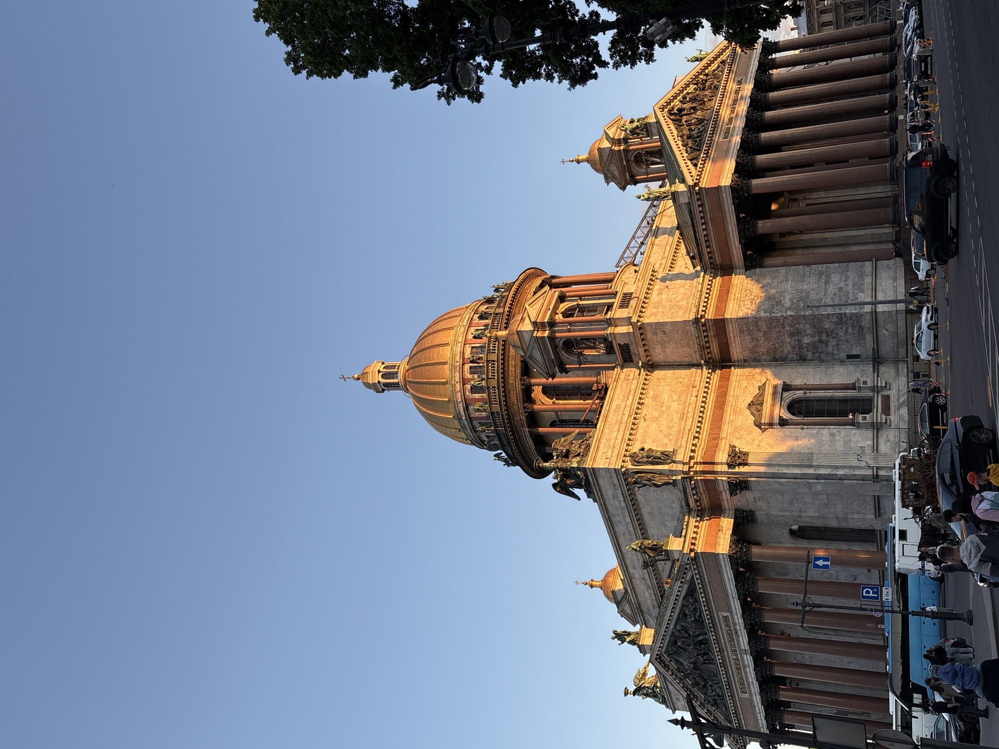 An exterior, low-angle, full shot captures St. Isaac's Cathedral in Saint Petersburg, Russia, under a clear blue sky, bathed in the warm, orange glow of late afternoon or early morning sunlight. The grand cathedral, a prominent landmark, features a large, golden dome topped with a cross and intricate architectural details, including multiple smaller domes, columns, statues, and a pediment adorned with sculptures. The sun illuminates the right side of the building, casting long shadows. In the foreground, a street scene unfolds with several people walking and gathered on the sidewalk. A bright blue bus, a horse-drawn carriage, and various parked and moving cars, including a black SUV, are visible on the road and street. A street lamp stands to the right of the frame, partially obscuring a dense green tree. The scene conveys a sense of historic grandeur and urban life.