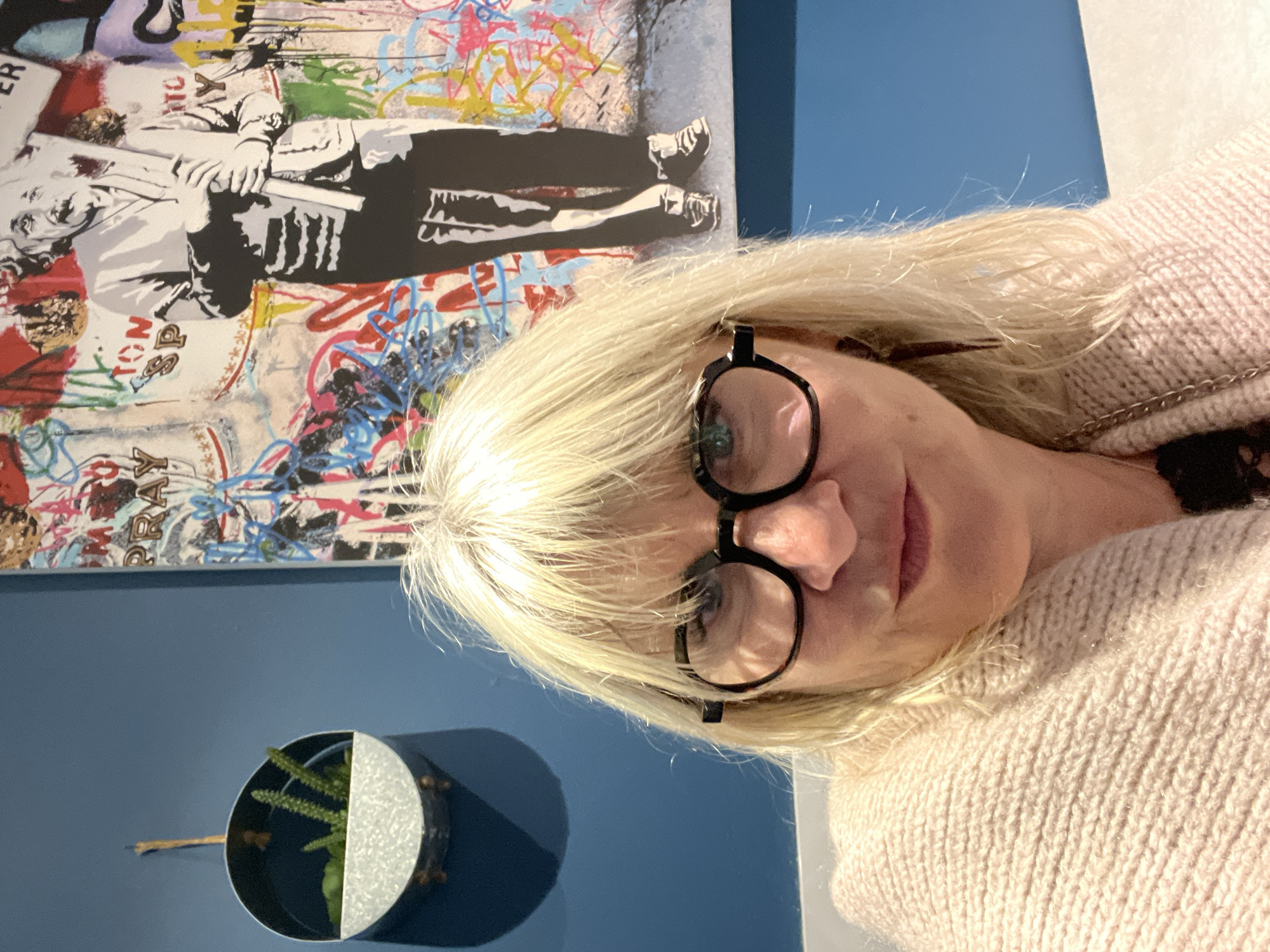 A medium, indoor shot depicts a blonde, light-skinned woman with fair features and glasses looking into the camera. She is wearing a light-colored knitted sweater, the texture of which is emphasized by the lighting. Behind her is a blue wall on which is mounted a metal, semicircular plant holder with a green plant inside. Above the planter is a large, abstract artwork. The artwork is vertically oriented and depicts a stylized silhouette of a person with a placard over a background of graffiti style colors and patterns. The setting is in Grimaud, France.