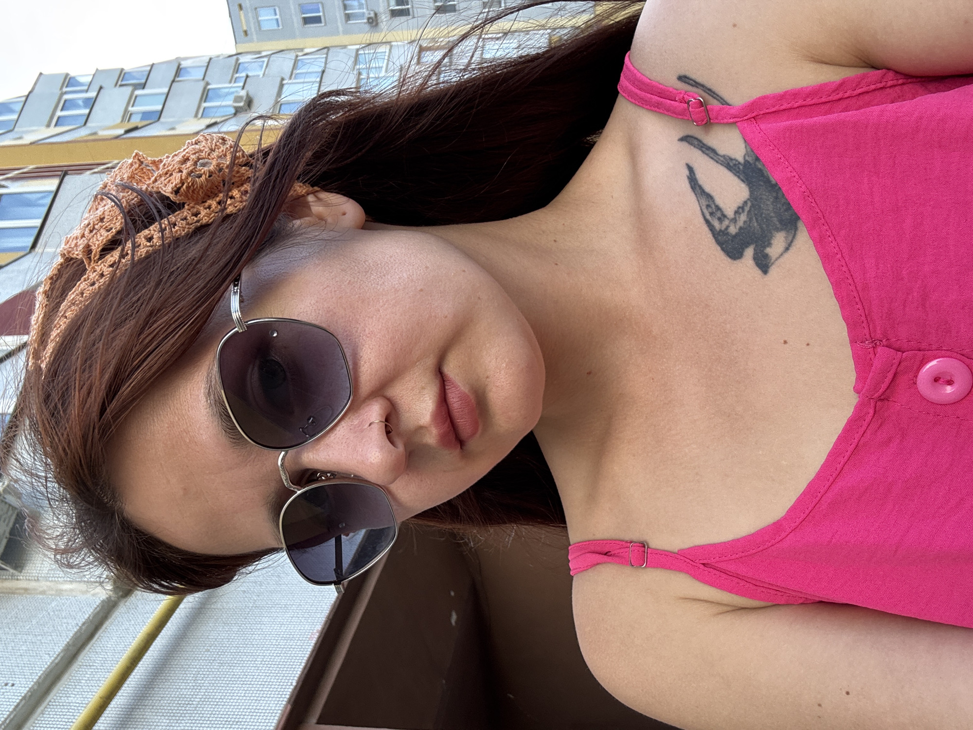 The image shows a medium close-up of a young woman with long, dark brown hair, wearing a bright pink sleeveless top and sunglasses. She has a knitted orange headband on her head and a small tattoo of a bird on her chest. The background appears to be an urban environment, with a building visible behind her. The scene is shot outdoors, possibly on a balcony or rooftop, given the presence of a railing. The woman is looking down and to her left. The specific location in the city of Naberezhnye Chelny, Russia cannot be determined from the image alone, as there are no identifying landmarks.