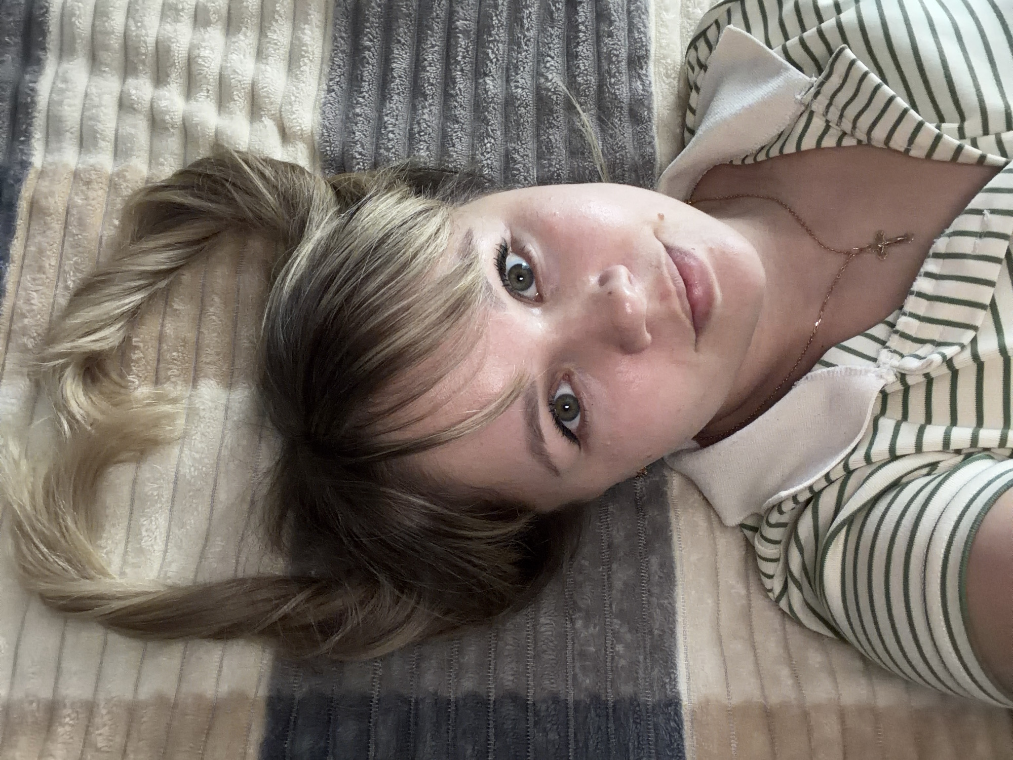 A young woman is depicted lying down, her head rotated to the left, looking directly at the camera with wide, green or hazel eyes. Her light blonde and brown hair is styled with bangs across her forehead and appears to be in two twisted or braided pigtails that fall to the left and right of her head. She has a subtle, engaging expression. She is wearing a collared top with alternating green and white horizontal stripes. A thin, gold-colored necklace with a small cross pendant hangs around her neck. The background consists of a soft, textured blanket or throw with prominent horizontal stripes in shades of grey, white, and beige/tan. The setting appears to be indoors, possibly on a bed or sofa. No specific time of day, weather, or location cues are discernible, and there is no visible text.