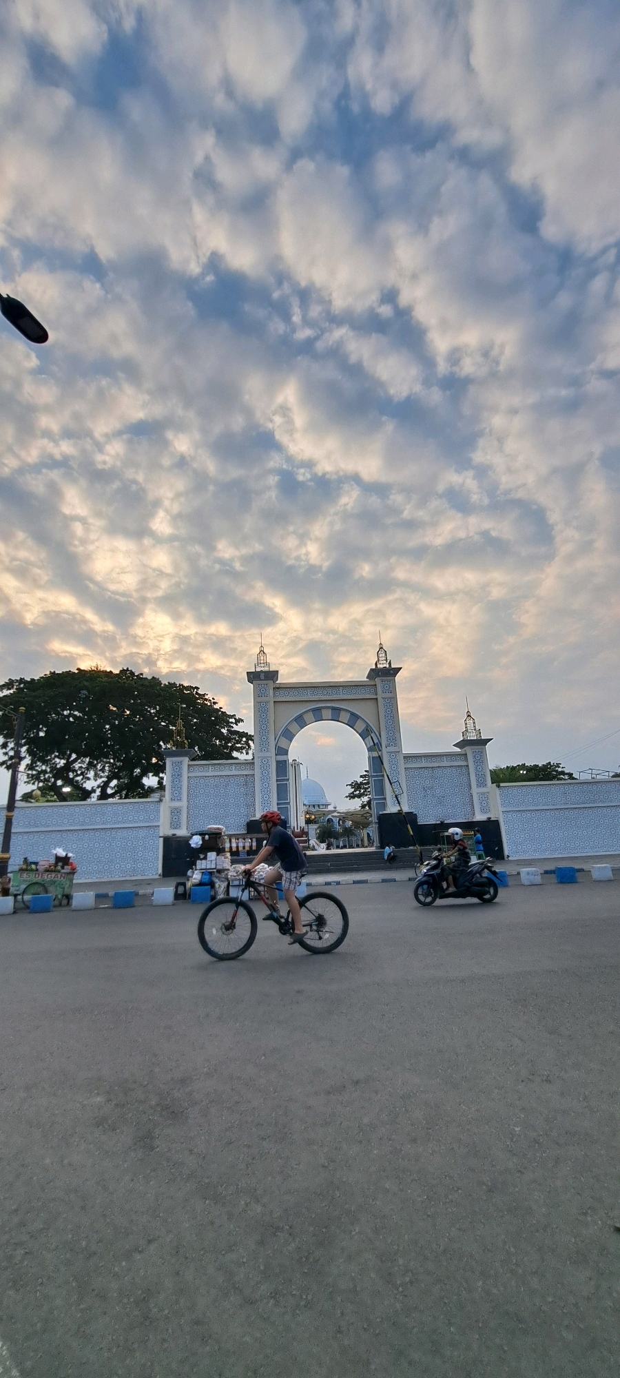 The image depicts a street scene in Pasuruan, Indonesia, with a prominent arched gateway in the background, likely an entrance to a significant building or complex, possibly a mosque given the dome visible through the arch. The sky is filled with scattered clouds, catching the warm light of what appears to be either sunrise or sunset. In the foreground, a person is riding a bicycle, wearing a red helmet and checkered shorts. To the right, another person is riding a motorcycle. The street is paved, with blue and white barriers lining the side. To the left of the gateway, there's a large tree and a blue and white patterned wall. A food cart is visible on the left side of the street. The overall atmosphere is one of everyday life in a city, with a blend of architectural elements and human activity under a dramatic sky.