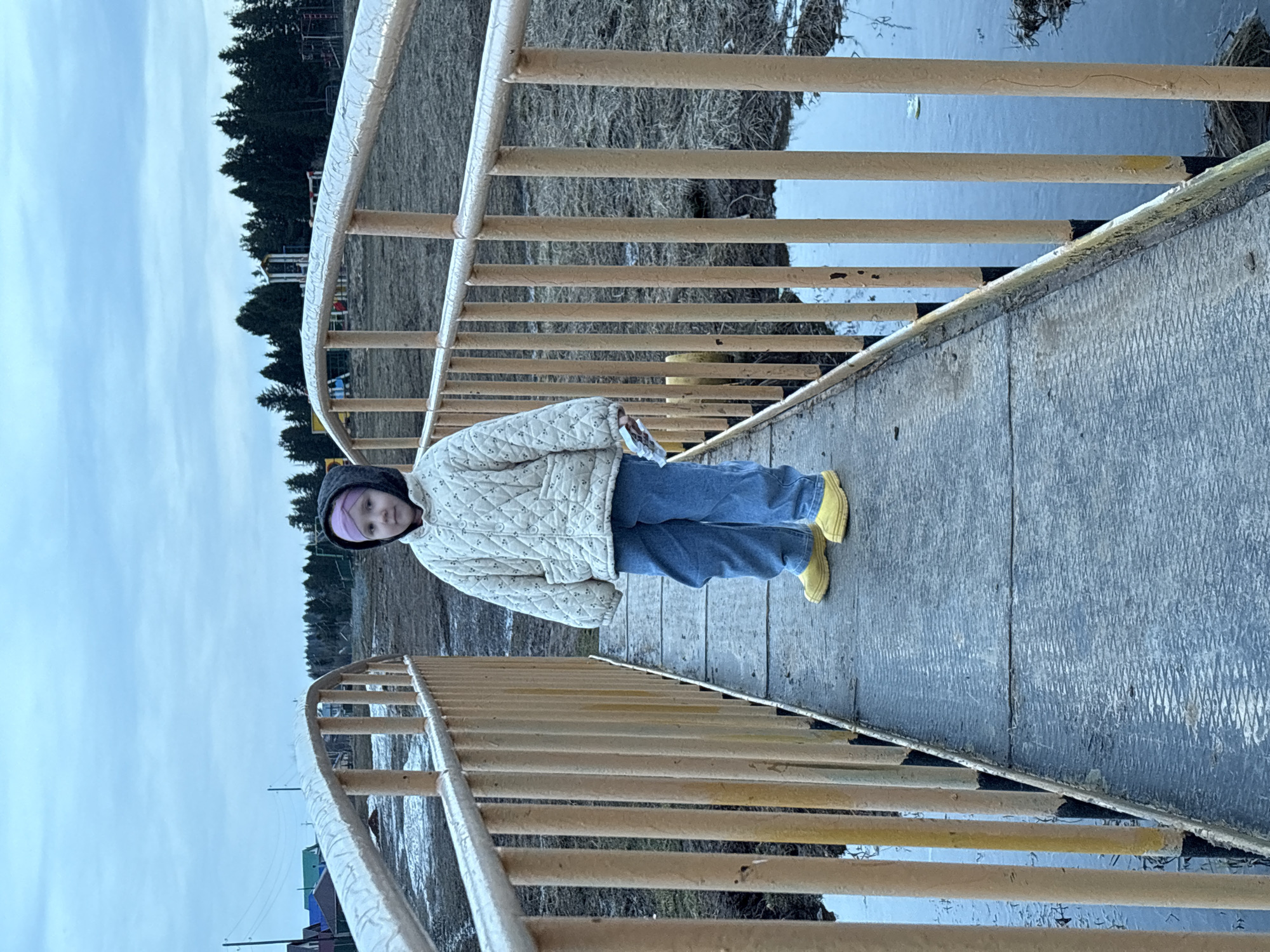 A young girl stands on a weathered bridge with yellow railings, likely in a park or outdoor area in Фоки, Russia. She is wearing a quilted white jacket, blue jeans, and bright yellow boots. She has on a dark head covering, possibly a hood or hat, with a lighter-colored band showing. The bridge appears to cross a waterway with ice or partially frozen water visible beneath it. The background includes trees, a gray sky, and hints of residential structures.