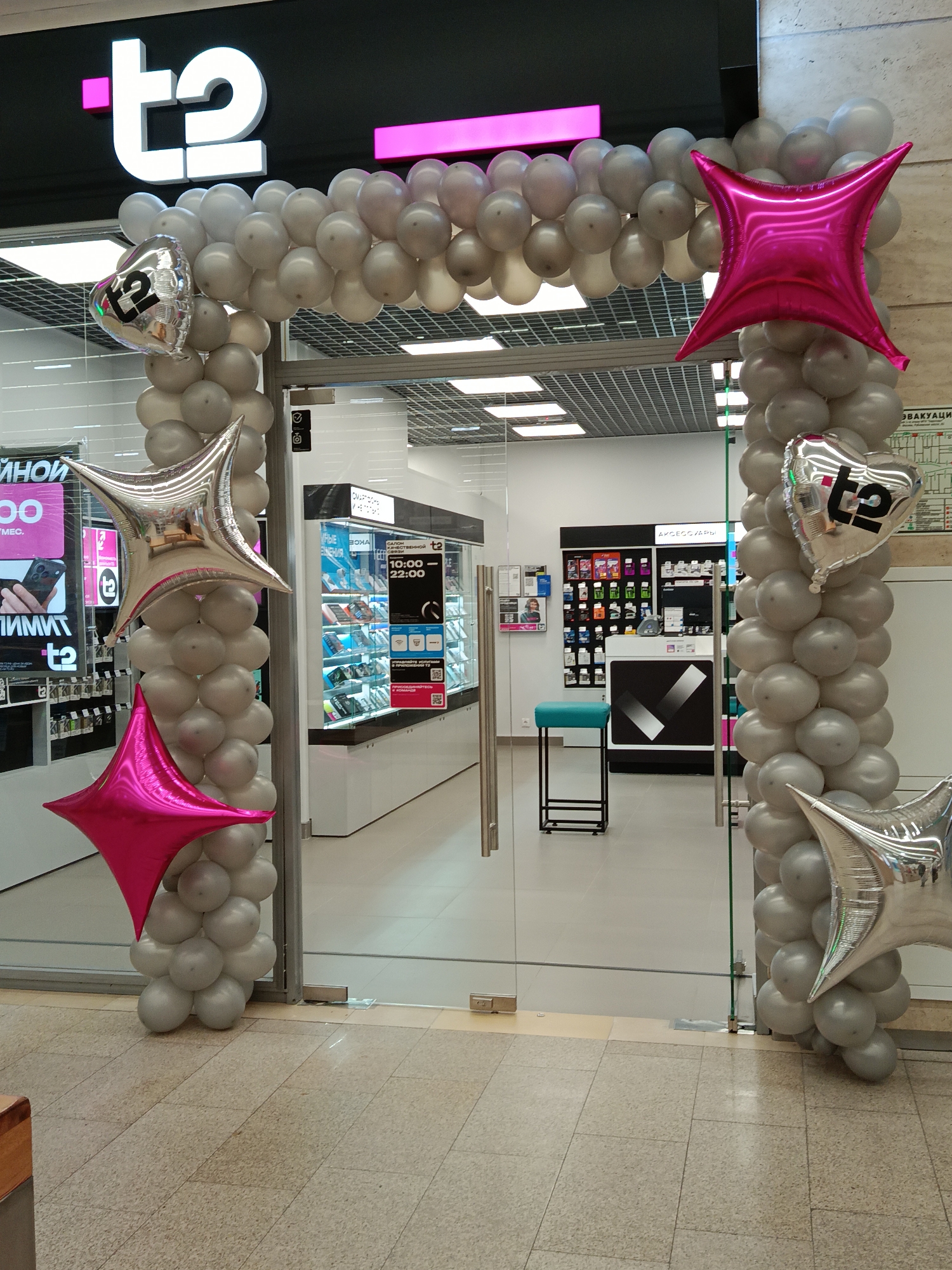 The image captures the entrance of a retail store, likely in a shopping mall, adorned with a festive balloon arch. The store is branded with a prominent "t2" logo in white and pink on a black facade above the entrance. A silver and grey balloon arch frames the clear glass double doors, featuring interspersed silver and pink star-shaped foil balloons. Two silver heart-shaped foil balloons, each also bearing the "t2" logo, are attached to the arch.
Inside the well-lit store, modern display cases showcase various electronic devices and accessories. A visible sign inside reads "АКСЕССУАРЫ" (Accessories), and another indicates operating hours as "10:00 - 22:00." A blue-seated stool is visible near a counter. The floor inside and immediately outside the store is tiled. On the right wall, a partial sign with the word "ЭВАКУАЦИЯ" (Evacuation) is visible. The scene suggests a recent grand opening or a special promotional event for the "t2" store, within an indoor commercial setting such as a mall in Moscow, Russia. No people are present in the image.
