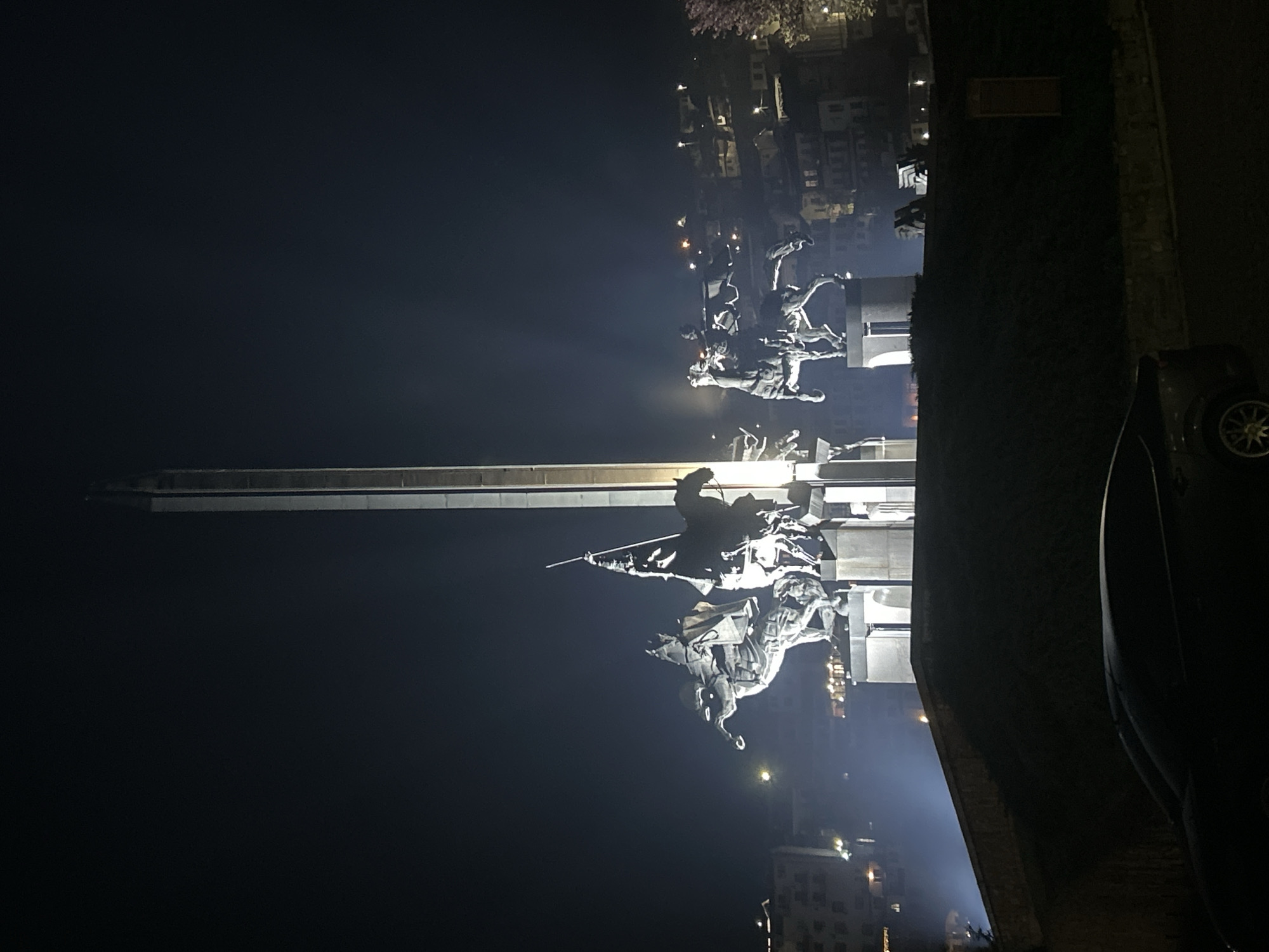 This image captures the Monument of the Asen Dynasty, a prominent landmark in Veliko Tarnovo, Bulgaria, at night. The monument, featuring dynamic sculptures of horsemen, stands against a dark sky and is illuminated by artificial lights. A tall, sword-shaped pillar rises behind the equestrian figures. In the background, the cityscape of Veliko Tarnovo is visible with scattered lights from buildings. A dark mass, likely vegetation and a partial view of a car, occupy the foreground on the right. The scene suggests a calm, possibly tourist-oriented, urban environment at night.