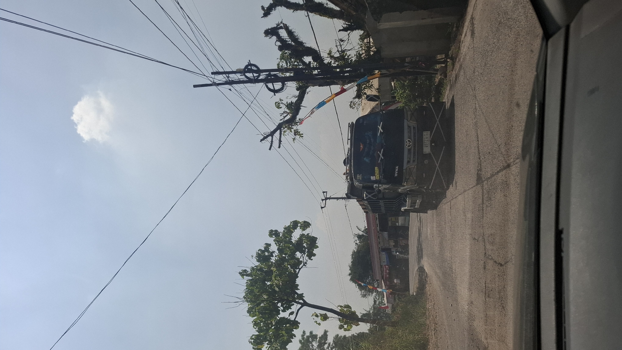 The image depicts a street scene in Cilaku, Indonesia, on a sunny day. In the foreground, the dashboard of a vehicle is visible, indicating the photo was taken from inside a car. The road ahead is paved and appears somewhat weathered. A black dump truck is the prominent subject, driving on the road and positioned slightly to the right of the center of the frame. The truck has a "DYNA" logo visible on its front.
To the left of the road, there is lush green vegetation, including a tree with a full canopy. Beyond the vegetation, a row of buildings with red roofs can be seen, suggesting a commercial or residential area. Several flags or banners in red, white, and blue are also visible along the street.
On the right side of the road, there is a concrete wall and a tall utility pole adorned with colorful bands, possibly part of a street decoration or warning system. There are numerous overhead power lines crisscrossing the sky, which is mostly clear with a single white cloud. The overall environment suggests a rural or semi-urban setting in Indonesia. There are no people clearly visible in the image, but the presence of the truck and buildings implies human activity.