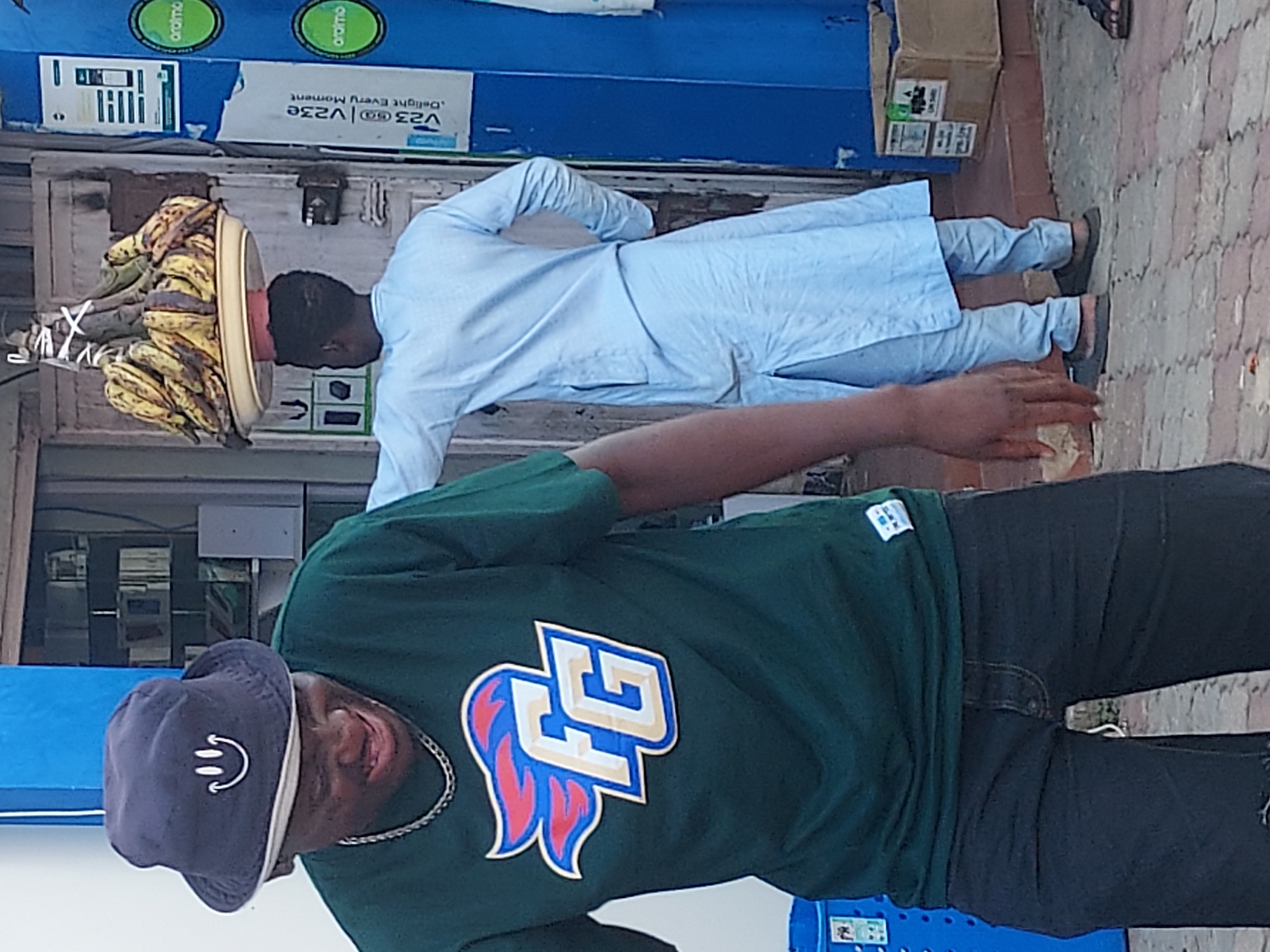 The image depicts two men on a street in Jos, Nigeria. In the foreground, a man wearing a dark green t-shirt with a stylized "FG" logo, dark jeans, and a dark blue bucket hat is partially visible from the torso down. He wears a silver chain around his neck. Behind him, a man is carrying a tray of plantains on his head. He wears a light blue, long-sleeved traditional garment and dark-colored shoes. The background includes a building with a blue sign that says "aramo" and "V23." The ground is paved with bricks.