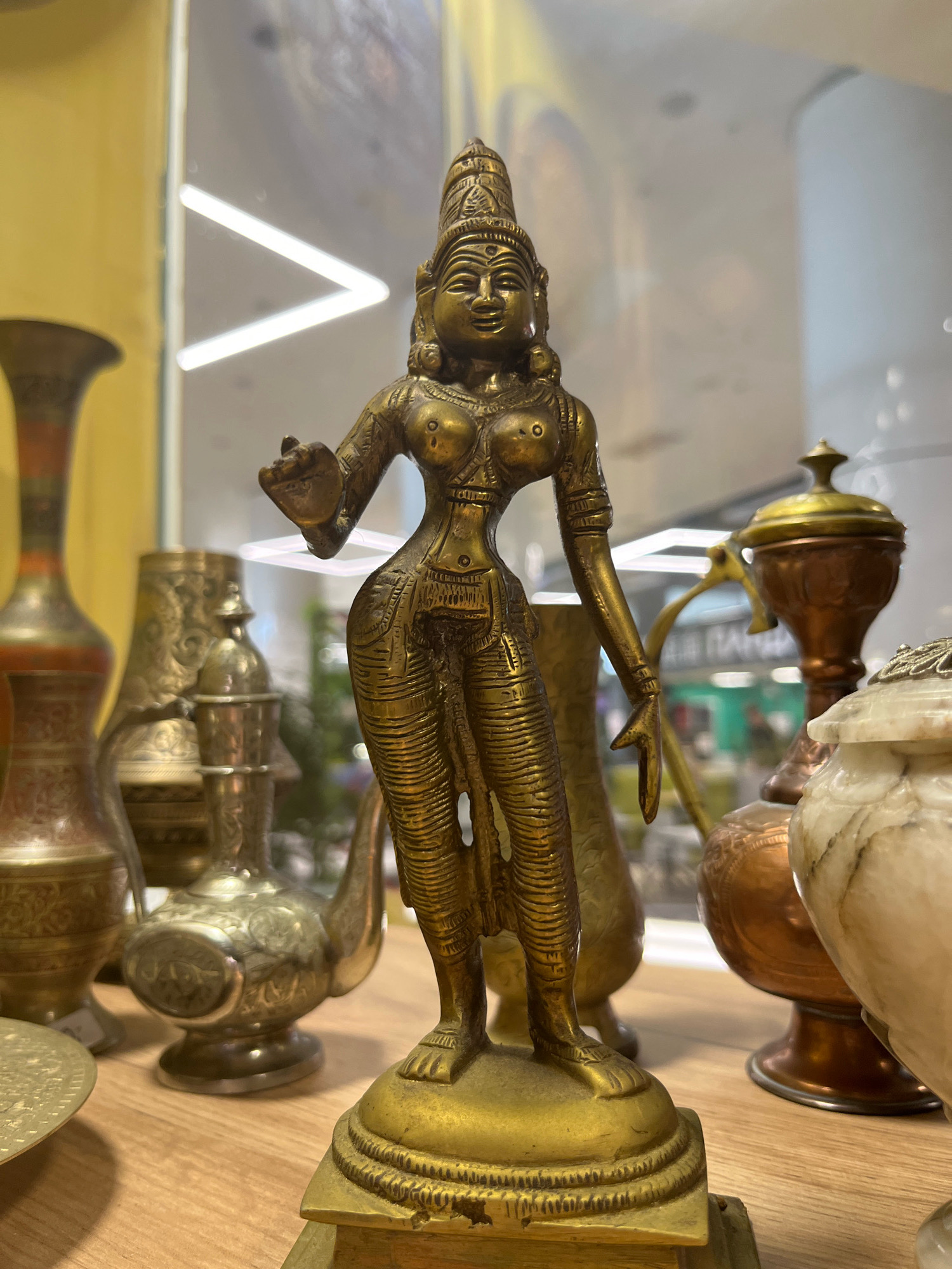 An ornate brass figurine of a woman stands in the center of the image, adorned with traditional Indian attire and jewelry. The figurine is placed on a wooden surface, possibly a shelf or display table. To the left and right of the figurine, various other decorative items are visible, including brass vases, a copper jug, and a marble container. The background suggests an indoor setting, possibly a shop or museum, with soft lighting and blurred reflections of white linear lights. The overall composition highlights the detailed craftsmanship of the brass figurine and its surrounding decorative objects.