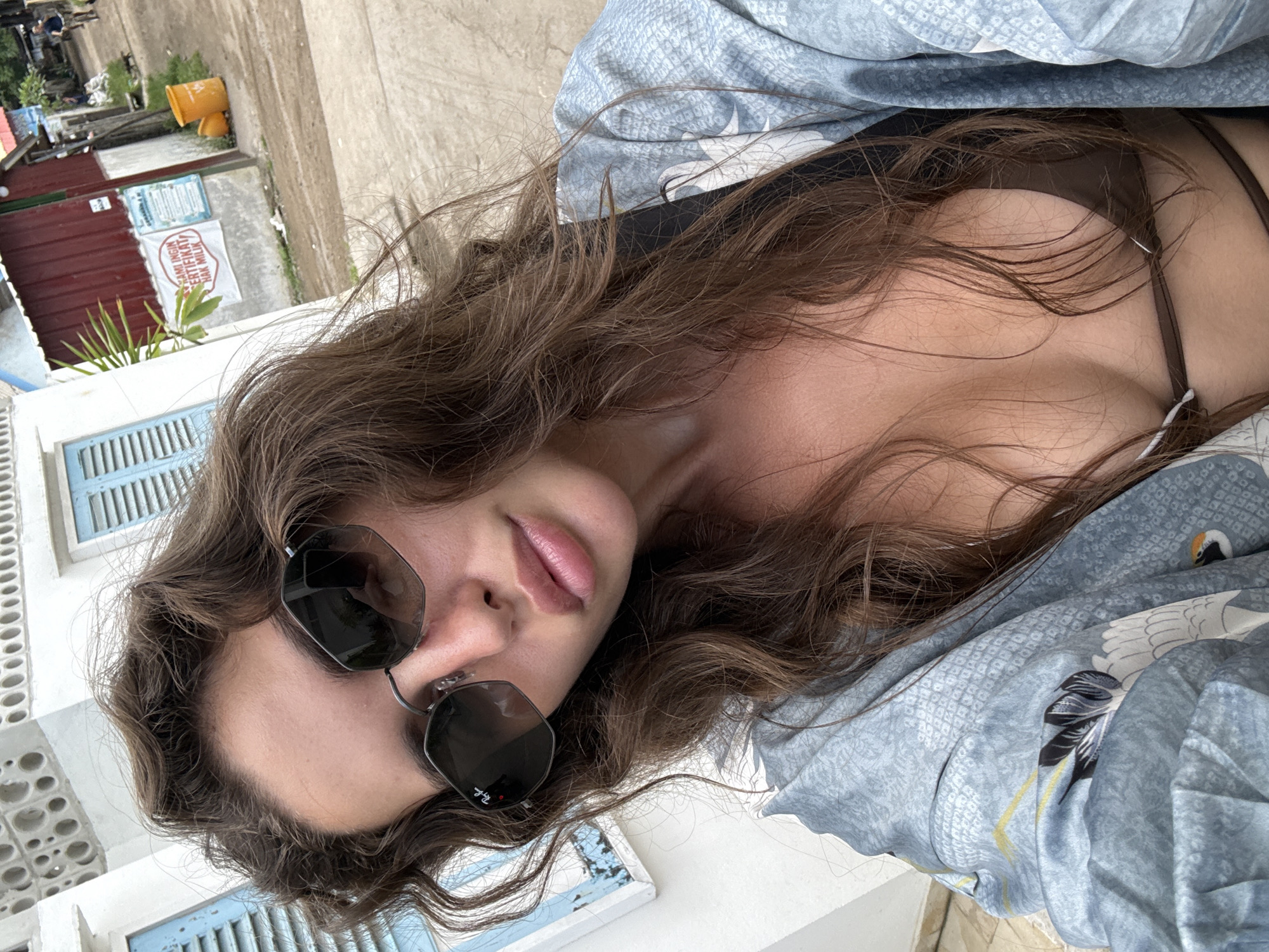 A woman wearing sunglasses is shown in a low-angle, portrait-style shot. She has wavy brown hair and is partially covered with a blue patterned garment. A brown swimsuit top is visible under the garment. Outside, the backdrop consists of a white building with blue shutters and a red metal structure with signs and foliage. A yellow object is also visible in the distance. The location is identified as Unknown, Indonesia.