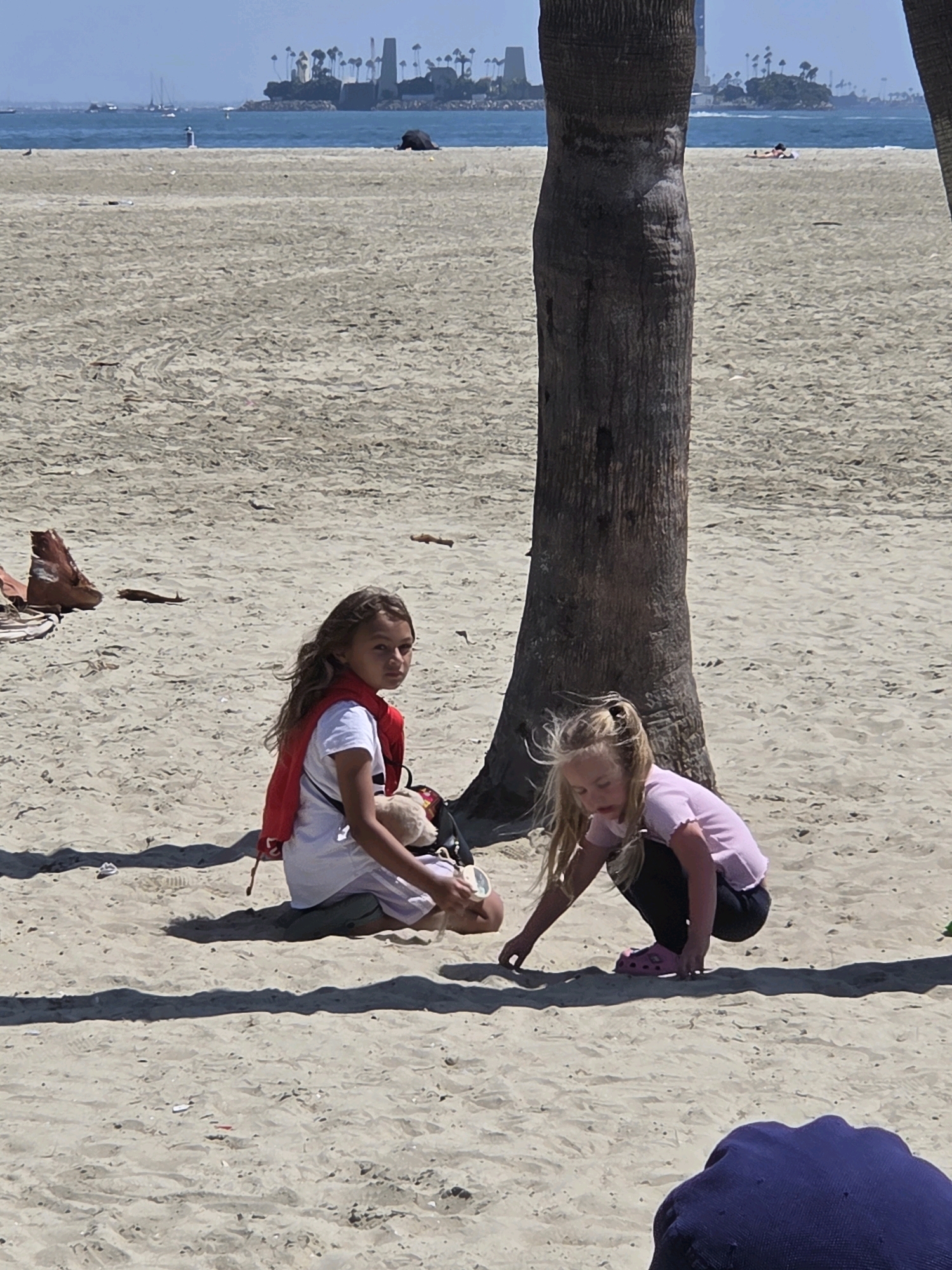 Two young girls are on a sandy beach in Long Beach, California, near a palm tree. The girl on the left, with long brown hair, wears a white t-shirt, a red vest, and dark shorts. She is seated and looking towards the camera. The girl on the right, with blonde hair, is crouching and appears to be playing in the sand. She wears a pink t-shirt and dark pants with pink Crocs. In the background, the ocean is visible with a distant shoreline featuring buildings and palm trees. The sand is light brown and appears somewhat rippled.