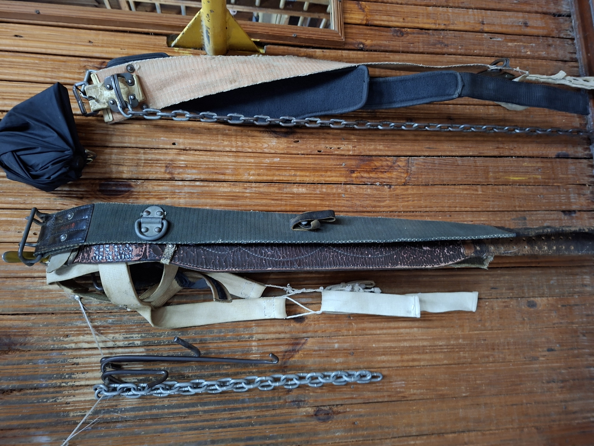 The image displays a collection of old, weathered equipment hanging on a wooden wall. On the left, a metal chain is suspended from a hook attached to a wire strung across the wall. To the right of the chain, a dark, curved metal object, possibly a hook, is also hanging. Further to the right, a wide, dark green strap with worn leather accents and metal fixtures is visible. Next to this, a lighter colored fabric strap with similar metal hardware is attached. Above these straps, a crumpled black item, resembling a bag or cloth, is hanging. On the far right, another wide strap, primarily beige with a black underside, is suspended. A metal chain hangs alongside this strap. The wooden wall has visible grain and appears to be old. There are no people or animals present in the image. The scene is static and appears to be an indoor storage area. There is no information to suggest this location is in Kazan, Russia.