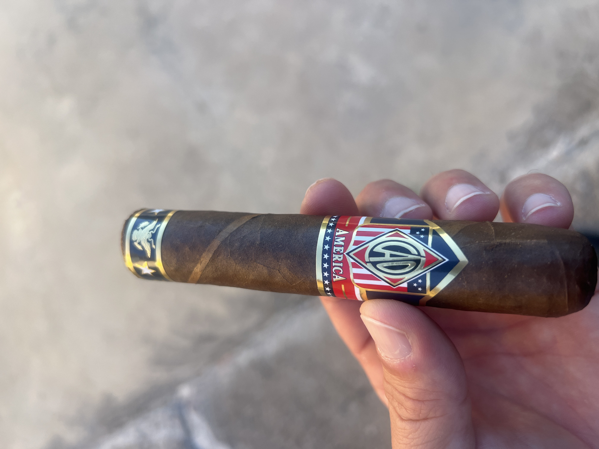 A person's hand is holding a cigar with a red, white, and blue band that says "AMERICA" and has stars and a logo with the letters "AD". The background is a blurry, light-colored surface.