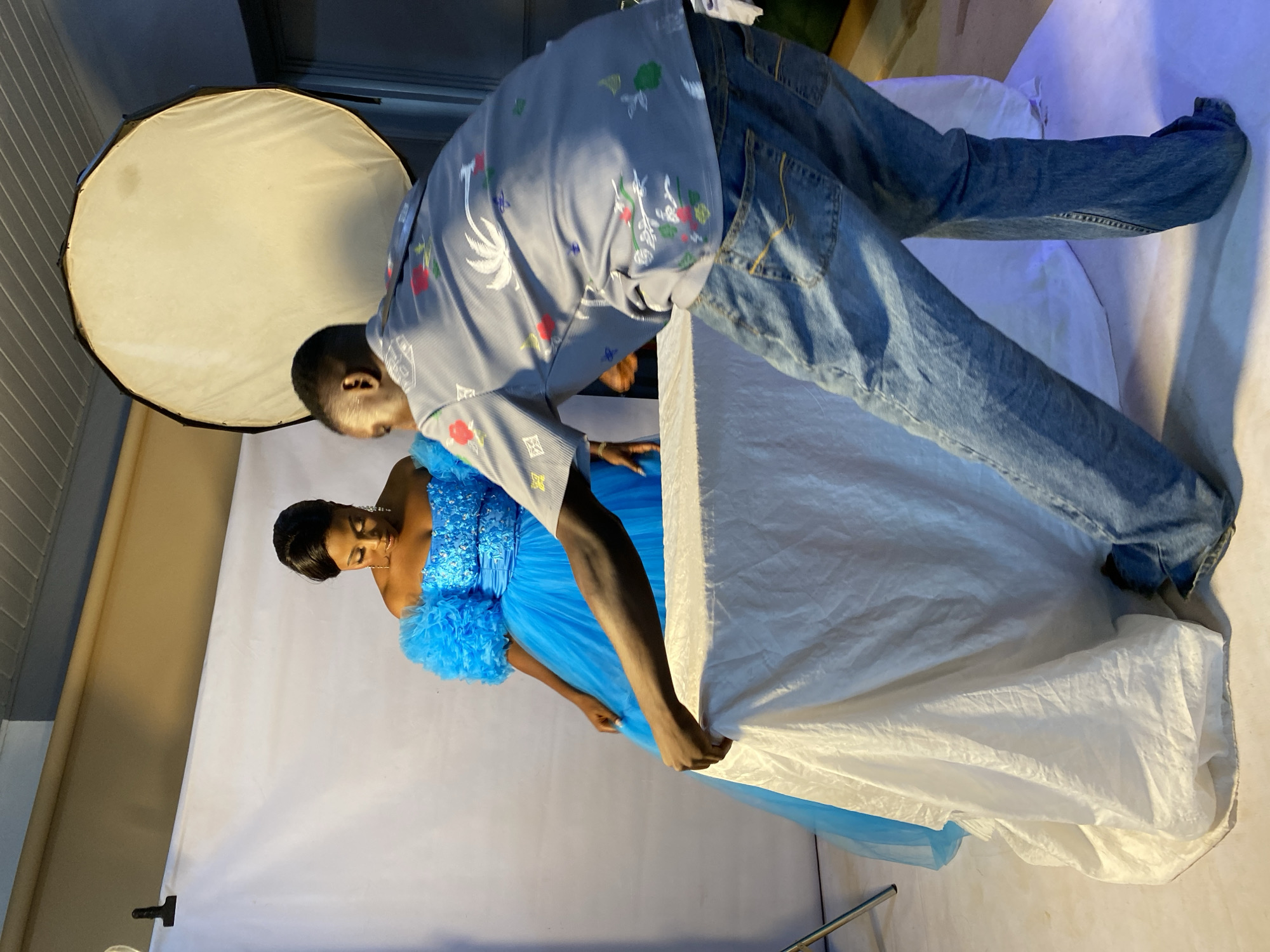 The image shows a photographer adjusting a large white fabric for a photoshoot. A woman in a vibrant blue, ruffled gown is positioned behind the fabric. The photographer, wearing a grey patterned shirt and blue jeans, leans over to manipulate the fabric, his hands visible. The woman is looking down, her expression unreadable. The scene appears to be an indoor studio setup with lighting equipment, including a large circular light diffuser on the left. The background consists of a light-colored wall and a roll of beige paper, possibly for backdrops. The overall lighting suggests an artificial studio environment, likely during daytime. The context of Bukuru, Nigeria, provides a geographical location for this photographic session.