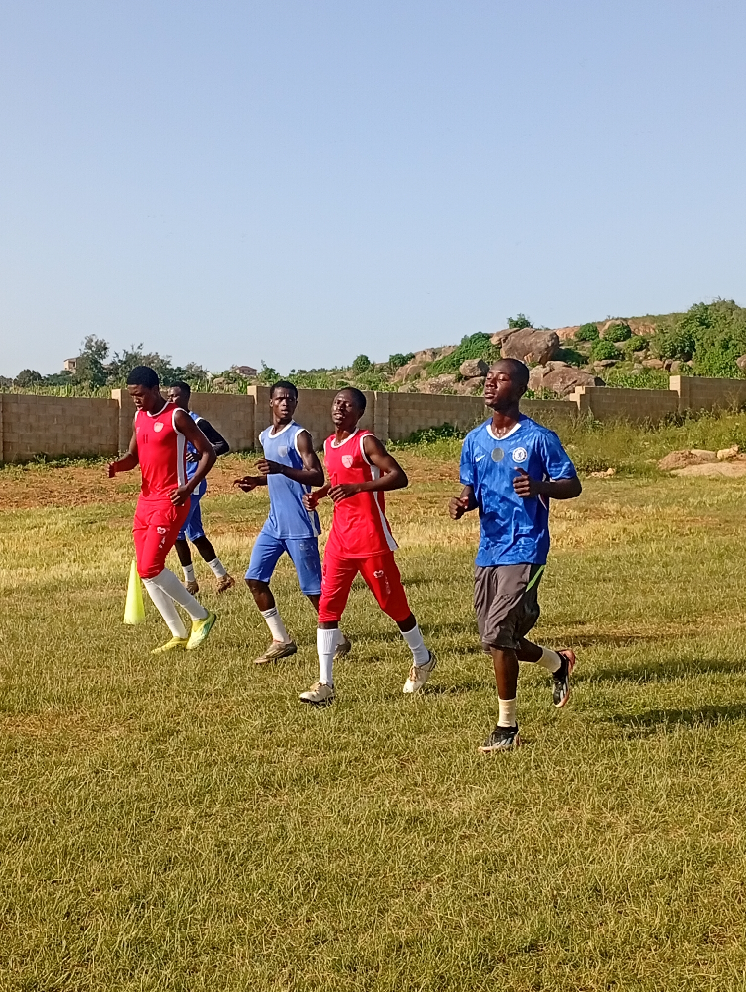 The image depicts a group of young men in athletic attire running on a grassy field. The individuals are dressed in a mix of red and blue jerseys, with some wearing shorts and athletic shoes. One individual in the foreground, wearing a blue jersey and gray shorts, is jogging with his eyes closed. Another person in a red jersey is partially visible on the left. In the background, a low brick wall runs horizontally across the scene, with a gently sloping, rocky, and vegetated hillside behind it. The sky is clear and blue. This scene is consistent with an outdoor sports training session, possibly at a training ground or a local park in Jos, Nigeria. The overall atmosphere is one of physical activity and training under a bright sky.
