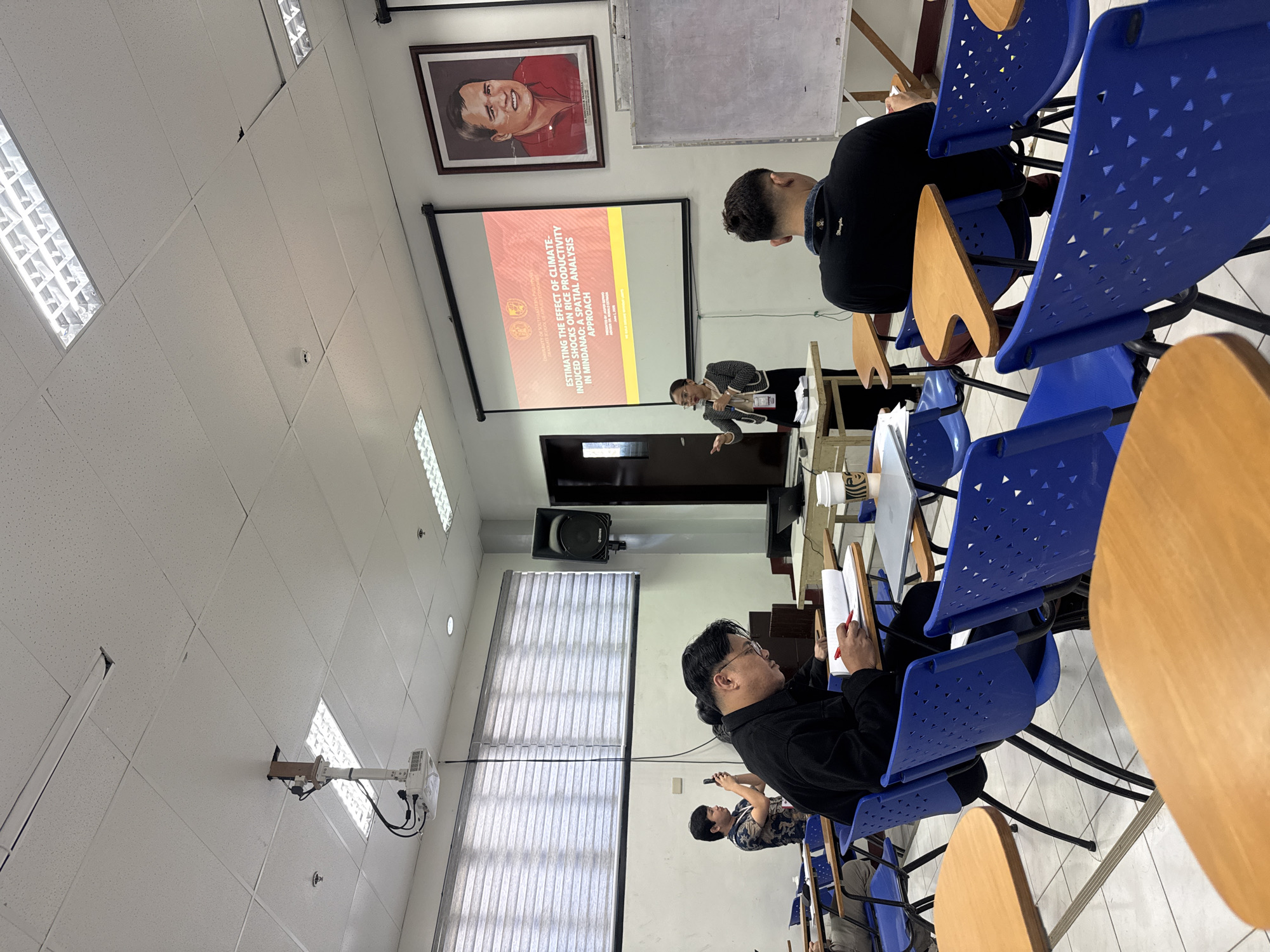 The image shows a classroom setting in Davao City, Philippines, where a presentation is being given. A woman is standing in front of a projection screen, speaking into a microphone. The projection screen displays the title of the presentation, "ESTIMATING THE EFFECT OF CLIMATE-INDUCED SHOCKS ON RICE PRODUCTIVITY IN MINDANAO: A SPATIAL ANALYSIS APPROACH." Several people are seated in the classroom, facing the presenter. One person in the foreground is taking notes. The classroom has blue plastic chairs with wooden writing surfaces. The walls are white with recessed lighting. A framed portrait hangs above the projection screen. A projector is mounted on the ceiling. There is a whiteboard next to the portrait.