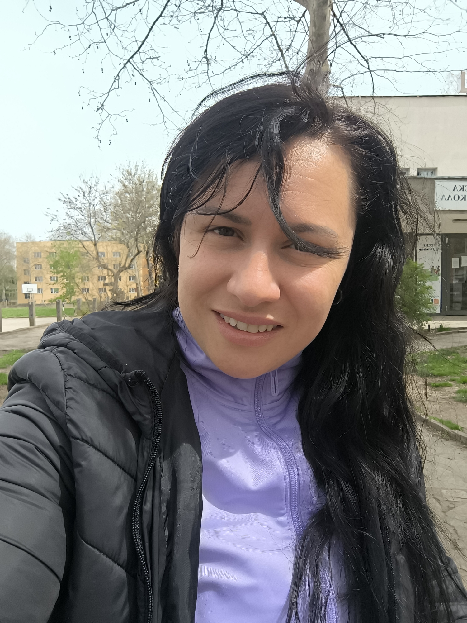 A medium, close-up shot depicts a dark-haired woman with wet hair, wearing a black puffer jacket over a lavender-colored athletic top, smiling at the camera while winking her left eye. The background reveals an urban environment, showing a basketball court, buildings, and trees. The location is identifiable as Pleven, Bulgaria, based on the Cyrillic script visible on a sign on the building behind her. The time of day appears to be daylight hours on a partly cloudy day, based on the sky and the amount of light.