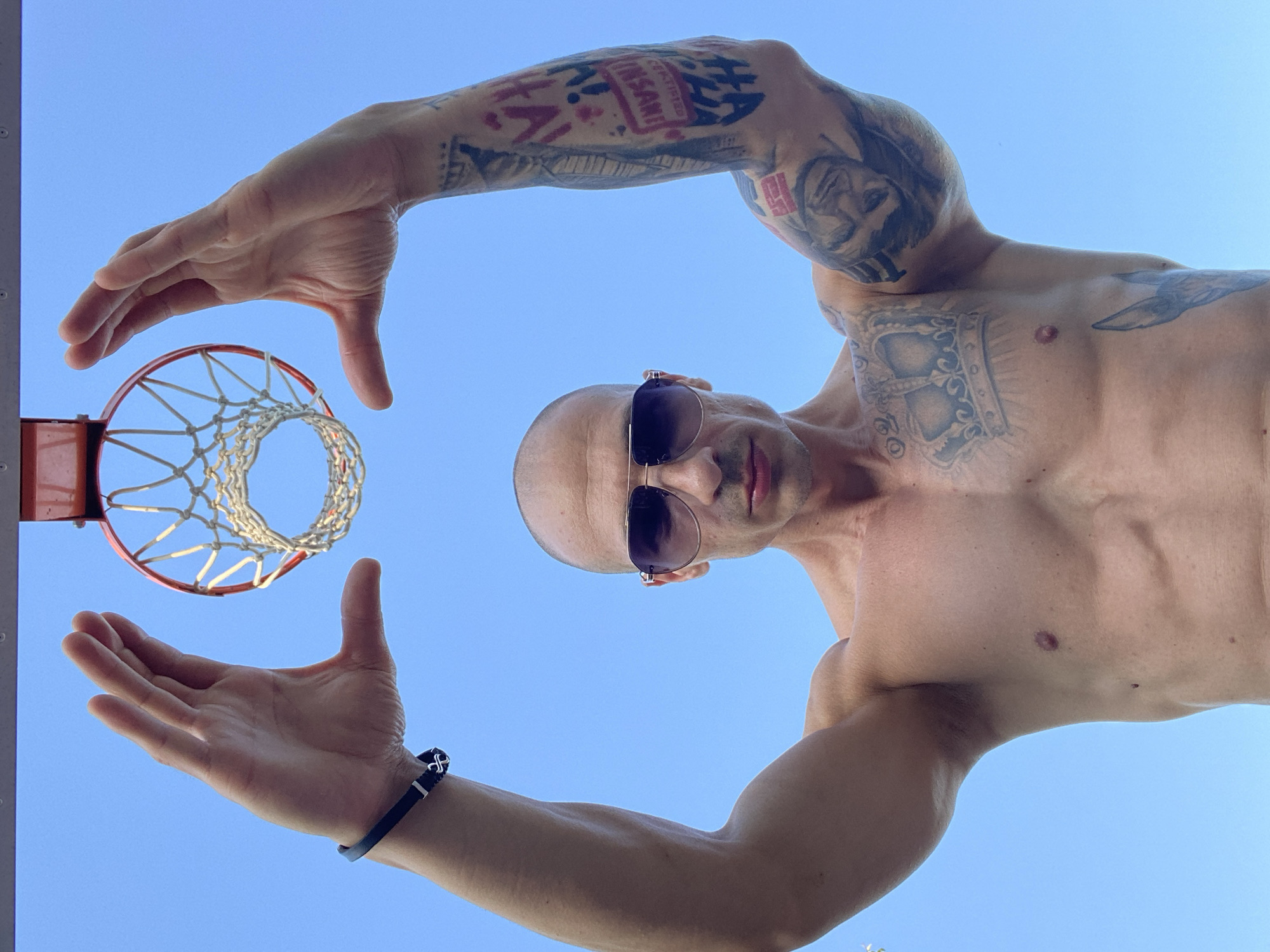 An outdoor, low-angle, medium shot captures a bald man with sunglasses, shirtless, against a clear blue sky. He is holding his hands up, framing a basketball hoop above his head. His left arm has a black bracelet. He has extensive tattoos on his right arm and chest, including a crown. The scene is set in Pleven, Bulgaria.