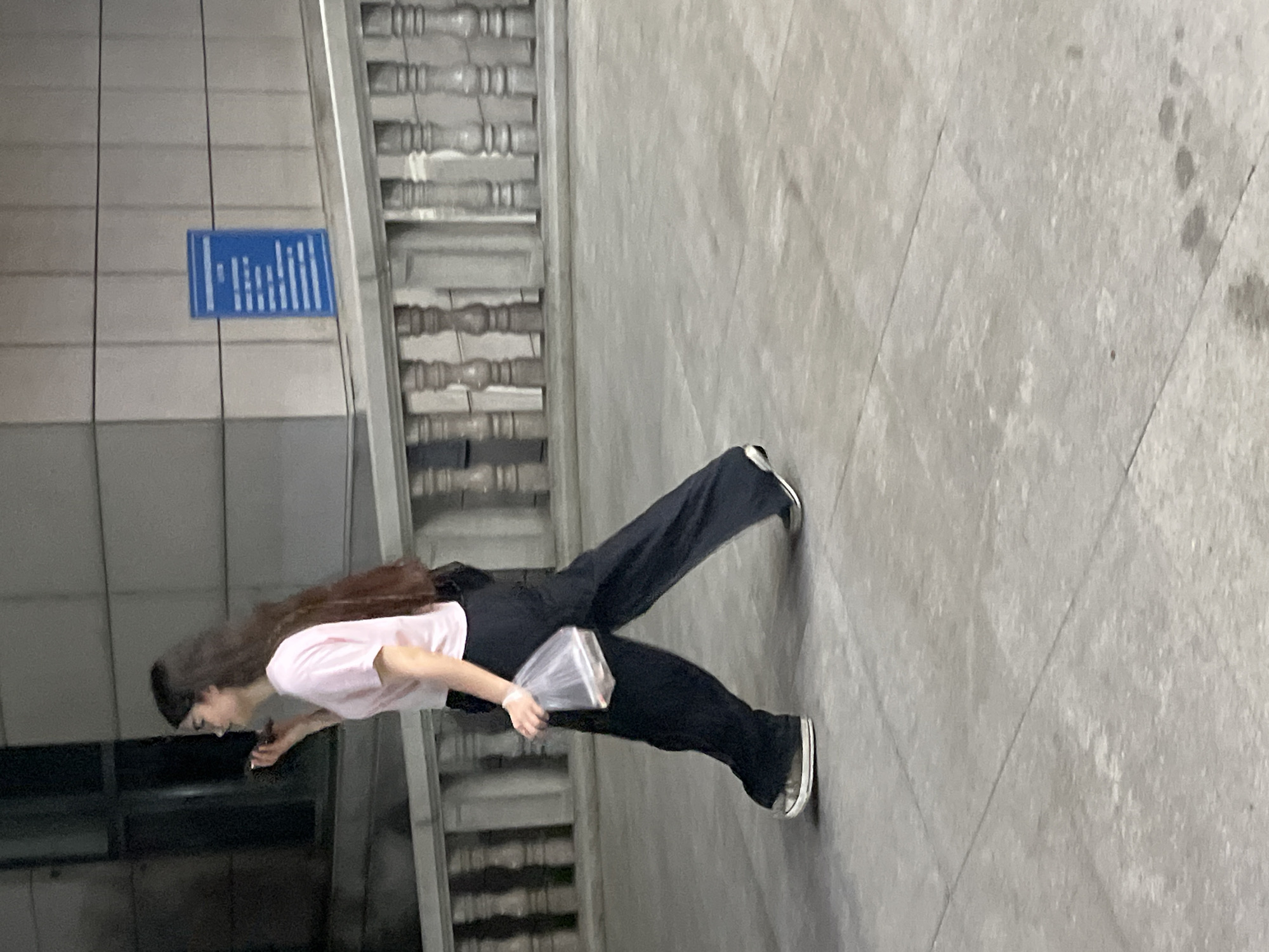 A medium shot shows a person with long brown hair and wearing a light pink t-shirt and black trousers walking on a stone-paved surface. The person is holding an object wrapped in a plastic bag in their right hand. Behind the person, a concrete wall features a blue sign and decorative metal railings. The image was taken in Changchun City, China.