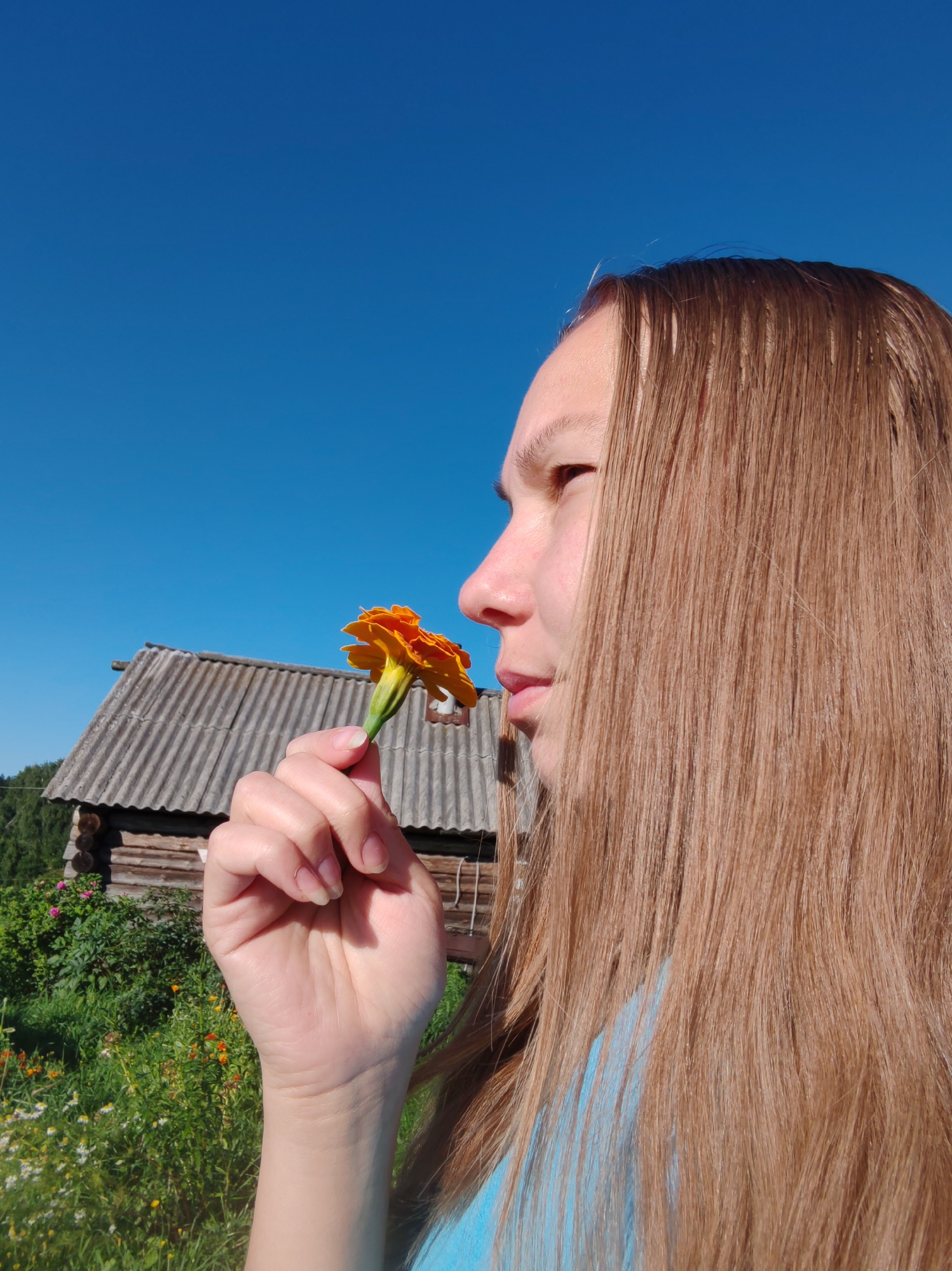 In an outdoor, rural setting, a young person with long, light brown hair is shown in profile, smelling an orange marigold flower held between their fingers. The person is wearing a blue top. Behind them, a rustic wooden building with a corrugated roof is visible, nestled amidst lush green vegetation and wildflowers. The sky is clear and blue, suggesting a sunny day. The overall scene evokes a sense of tranquility and connection with nature. The location is identified as Чухлэм, Russia.