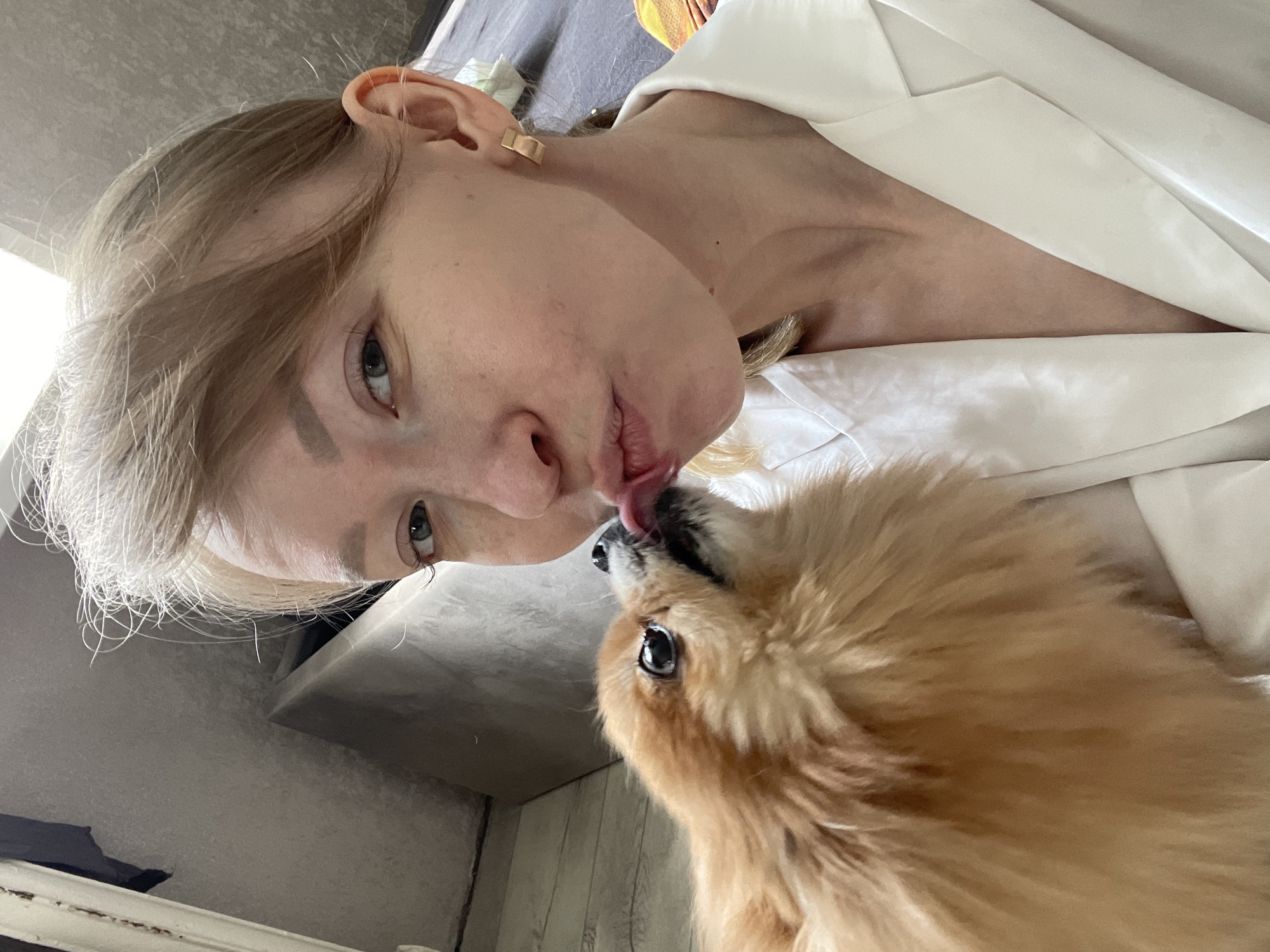 A woman with light skin and light brown hair is shown close-up in an indoor setting. She is wearing a white shirt or jacket. The woman is positioned close to a small, brown, fluffy dog. The dog is licking the woman's face near her mouth. A gray wall is visible in the background. The location is identified as Perm, Russia, as indicated by geotags or other location-based information.