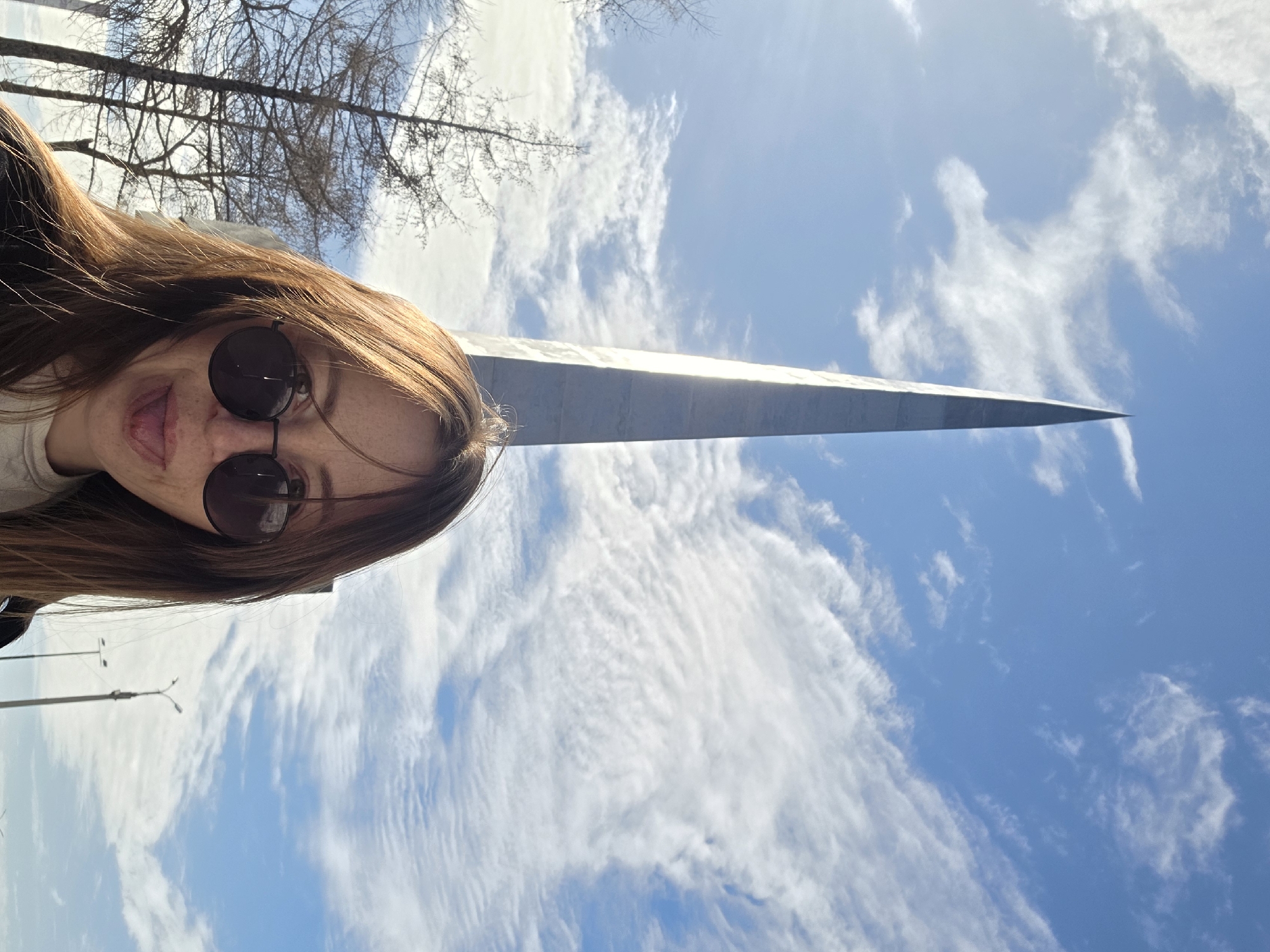 An outdoor photograph captures a female individual in the foreground, facing the camera and partially obscuring the lower section of a monument. She has long, light brown hair, wears round, dark-tinted sunglasses, and a light-colored top under a dark outer garment; her tongue is extended. Dominating the background is a tall, slender, grey obelisk with a sharp, pointed apex, identifiable as the Obelisk of Glory (Monument to the Fighters of the Revolution) in Votkinsk, Russia. The monument's right side is illuminated by direct sunlight. To the left, several bare deciduous trees are visible. The sky is bright blue with scattered white, cirrus-like clouds. In the far-right background, utility poles or streetlights are faintly discernible.