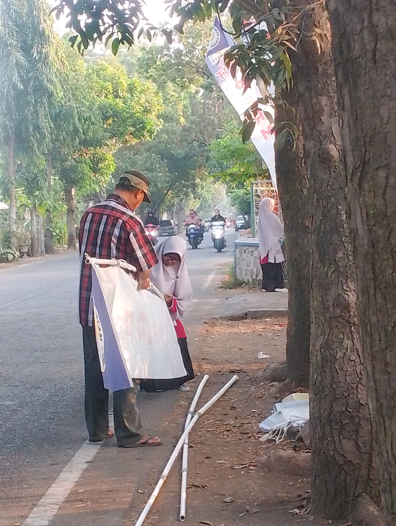 An outdoor, eye-level, medium shot shows a man and a young girl on the side of a paved road in what appears to be the city of Probolinggo, Indonesia. The man, wearing a plaid shirt and a cap, is facing away from the camera and holding a large white banner with blue and purple accents. The girl, wearing a light-colored hijab and red and black clothing, is facing the banner and appears to be interacting with it. Two long white poles, likely part of a flag or banner assembly, lie on the ground next to them. In the background, several motorcycles and a car are visible on the road, along with trees and foliage. The lighting suggests it is either early morning or late afternoon.
