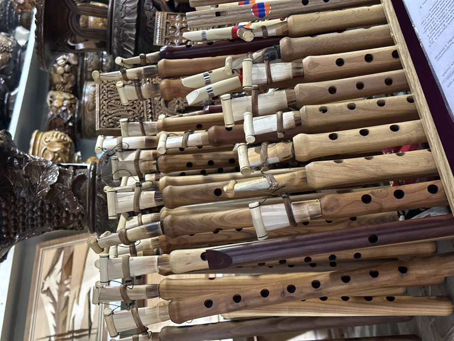 The image displays a dense collection of traditional Armenian duduks, presented upright in what appears to be a craft shop or market stall in Yerevan, Armenia. The duduks are primarily made of wood, exhibiting various shades from light tan to rich reddish-brown, and each features multiple circular finger holes along its length. At the top of each instrument is a light-colored cane or bamboo reed assembly, secured with string or twine. They are packed tightly into a wooden display rack, partially visible at the bottom right, which also shows a piece of white paper with Armenian text. In the background, out of focus, additional handcrafted wooden items are visible, including a framed wooden inlay artwork depicting a mountain landscape and intricately carved wooden vessels and boxes, some adorned with grape vine motifs. One duduk on the right side of the frame features a small ribbon displaying the red, blue, and orange colors of the Armenian national flag. No individuals are present in the scene.