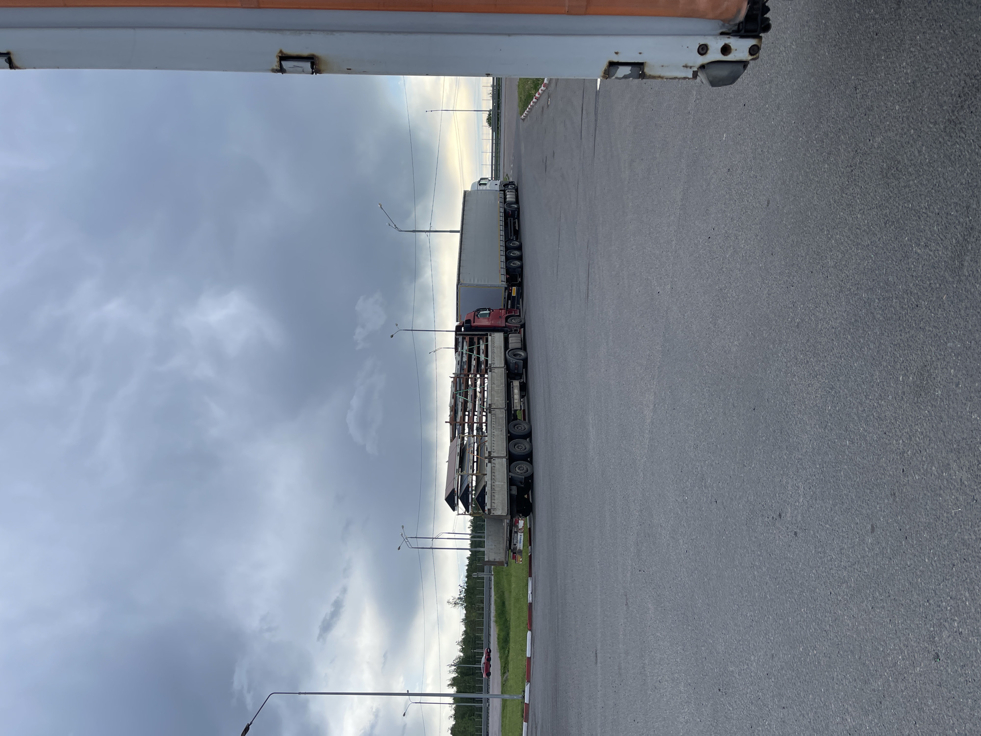 The scene shows two semi-trailer trucks parked on an asphalt road near what appears to be the edge of the city of Бавыкино, Russia. One truck is loaded with a metal construction, while the other has a white cargo container. Street lamps are visible along the roadside. The sky is cloudy. The image seems to be taken from a doorway or beneath a structure that obscures the upper left and right corners of the frame. There are no visible people in the scene.
