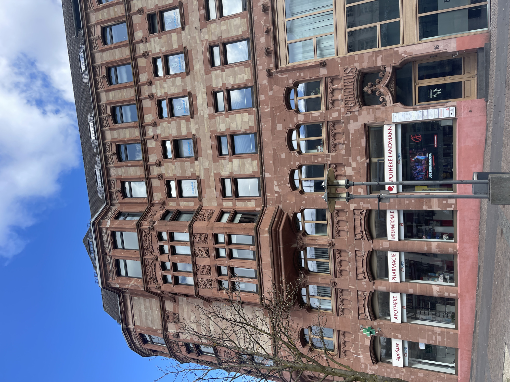 The image shows a tall, ornate red brick building in Saarbrücken, Germany. The building features multiple windows on each floor, some with decorative stone framing. Signage indicates the presence of a pharmacy, specifically ApoSaar, Apotheke, Pharmacie (AVIE), and Internationale, and another business, "POTHEKE LANDMANN". Above these businesses, the building’s name "TERMINUS" is displayed. A single tree with bare branches is visible at the bottom of the image, and the sky is a clear blue with a few scattered clouds. A traffic light pole and possible sidewalk are in the lower right. Some windows have signage for "H-Dental Zahnarztpraxis". The building is number 16 on the street.