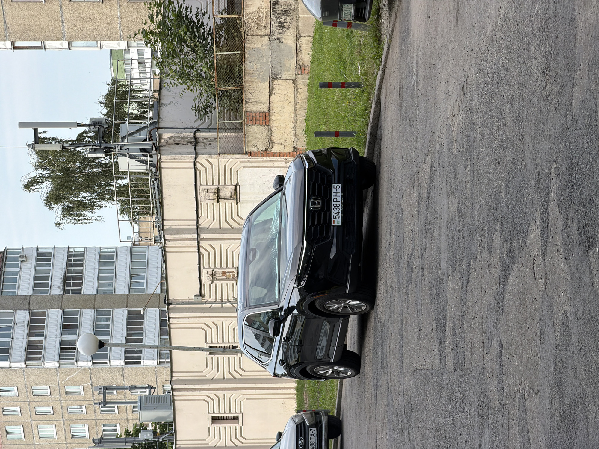 In Minsk, Belarus, a black 2023 Honda CR-V is parked on an asphalt road. The vehicle, facing right, has a license plate that reads "5438 PH-5". To its left, the front of another car is visible, displaying a license plate with "488 TA-7". In the background, a residential building with multiple stories and numerous windows stands tall. The environment includes a concrete wall with decorative patterns, a metal fence, and a patch of grass. Two vertical bollards with red and grey markings are positioned near the grass. The sky is overcast.