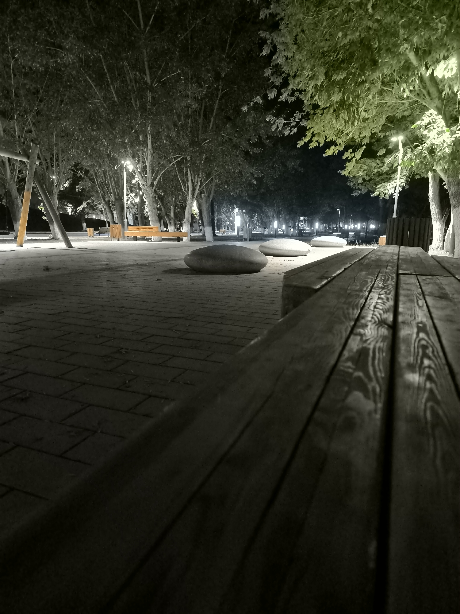 The image depicts a park at night in the city of Jaña Būqtyrma, Kazakhstan. The foreground features a close-up view of a wooden picnic table with a blurred perspective, highlighting the texture of the wood. Beyond the table, a paved walkway made of interlocking bricks is visible, illuminated by streetlights. Several large, smooth, oval-shaped stones are placed on the walkway. In the background, a row of trees lines the park, their branches reaching towards the sky. Wooden benches and what appear to be swings are also present. The park is sparsely populated, with no individuals clearly visible. The lighting suggests an artificial illumination system, likely street lamps, which cast a soft glow on the scene, creating a tranquil atmosphere.