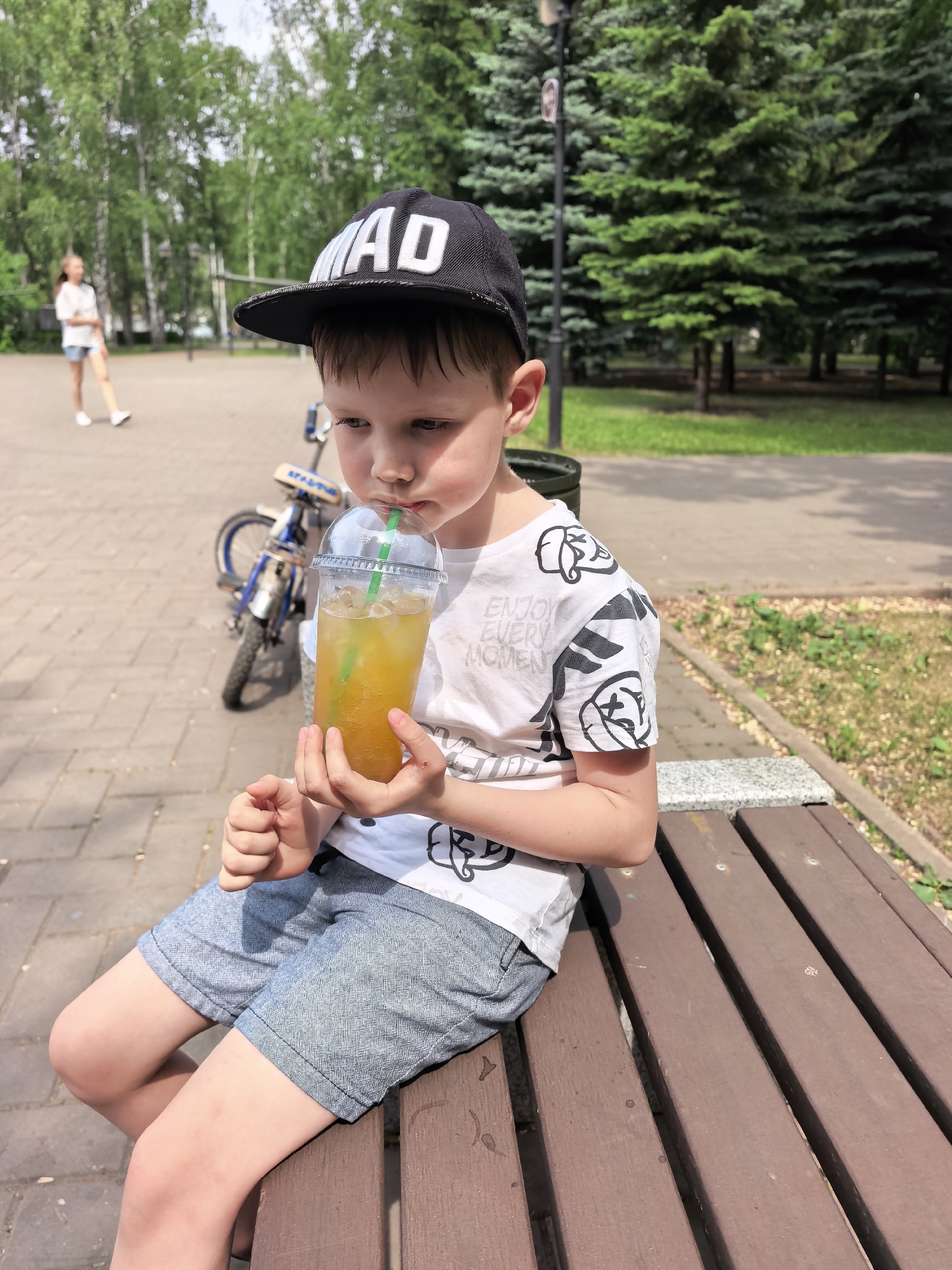 A young boy is sitting on a wooden bench in a park, holding a clear plastic cup filled with a yellow beverage and a green straw. He is wearing a black baseball cap with white lettering "MAD" on the front, a white t-shirt with black patterned designs, and blue shorts. He appears to be drinking from the cup. A bicycle is leaning against the bench behind him. In the background, there are trees, a paved walkway, and another person walking in the distance. Based on visual information and available details, the location is likely within a park in the city of Sterlitamak, Russia.